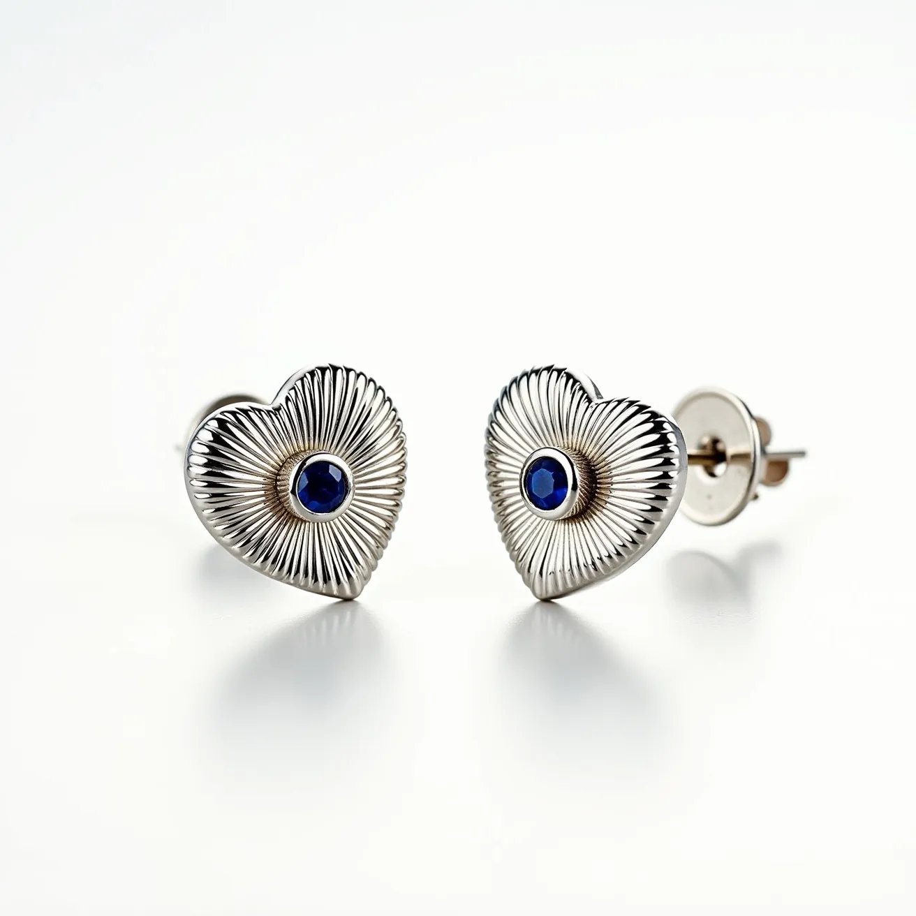 These heart earrings are crafted from a polished metal with a ribbed design that enhances their radiant appeal. At the center of each heart, a blue, round-cut gem is set, likely a sapphire, which adds a touch of elegance and color. The gemstone is securely held in place by a bezel setting, providing durability and style. The earrings are equipped with a post and butterfly clasp for secure and comfortable wear.