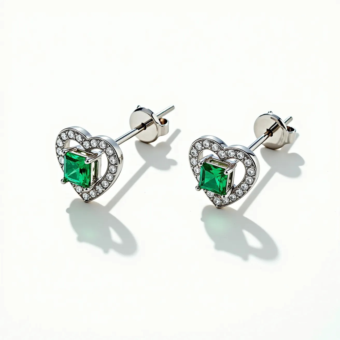 These heart earrings feature a sophisticated design characterized by a heart-shaped setting adorned with numerous small clear stones, likely diamonds, that accentuate the outer edge. At the heart's center, a prominent square-cut green gemstone, possibly an emerald, is prominently displayed, secured in a four-prong setting. The metal appears to be a polished silver or white gold, offering a sleek and shiny look to the overall composition. The earrings are equipped with a post and butterfly clutch back, ensuring a secure and comfortable attachment when worn.