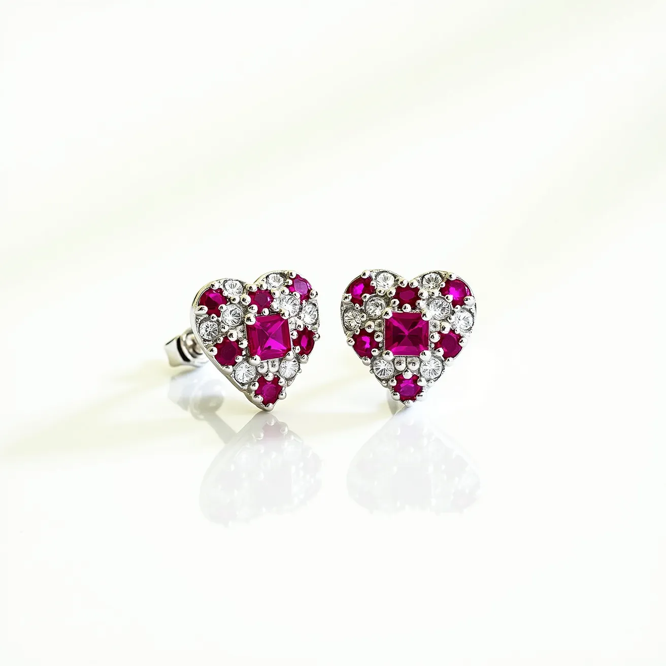 These heart earrings feature a striking design where each earring is crafted in a heart shape, composed of vibrant red stones and clear gems. The red stones, possibly rubies or garnets, are cut into square shapes at the center, surrounded by smaller, round clear stones, likely diamonds or crystal, creating a contrasting and luxurious effect. The stones are set in a metal that appears to be white gold or silver, enhancing the brilliance of the gems. Each earring is equipped with a post and butterfly clasp for secure wear, ensuring they remain comfortably in place. The arrangement of the stones within the heart design provides an elegant and timeless aesthetic.