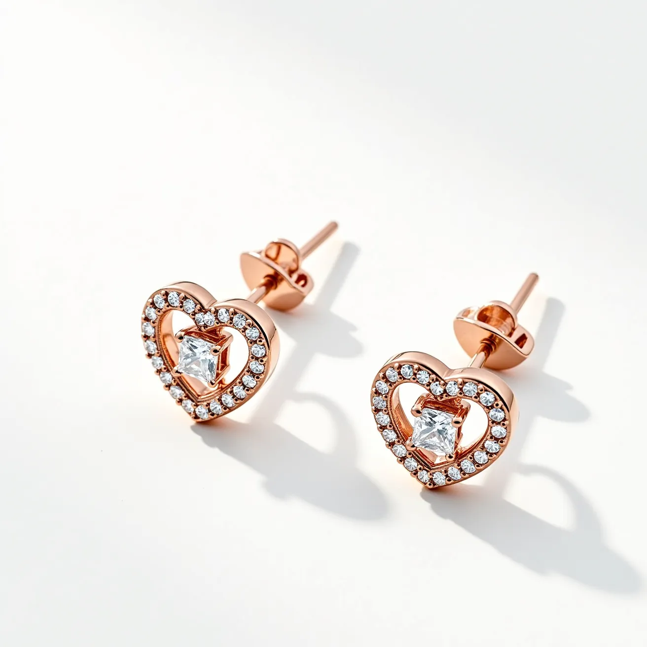 These heart earrings feature a rose gold metal setting shaped like a heart, intricately decorated with small, round-cut diamonds or diamond-like stones that encircle the outer edge. At the center of each earring is a square-cut clear gemstone, held securely in a prong setting. The design showcases a classic and elegant style, with the gemstones adding a sparkle that enhances the overall allure. These earrings are equipped with a standard post and butterfly back closure, ensuring a secure and comfortable fit when worn.