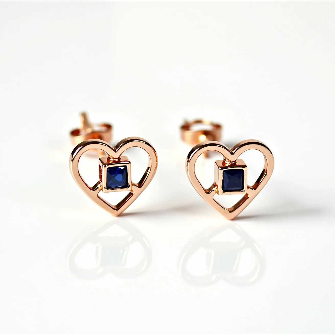 These heart earrings are crafted with a gold-colored metal forming an elegant open-heart design. Each earring features a square-cut blue gemstone set in a bezel setting at the center, adding a rich contrast to the metallic hue. The earrings are secured with a classic post and butterfly back clasp, ensuring stability and comfort for the wearer. The design combines simplicity and charm, making for a versatile accessory.