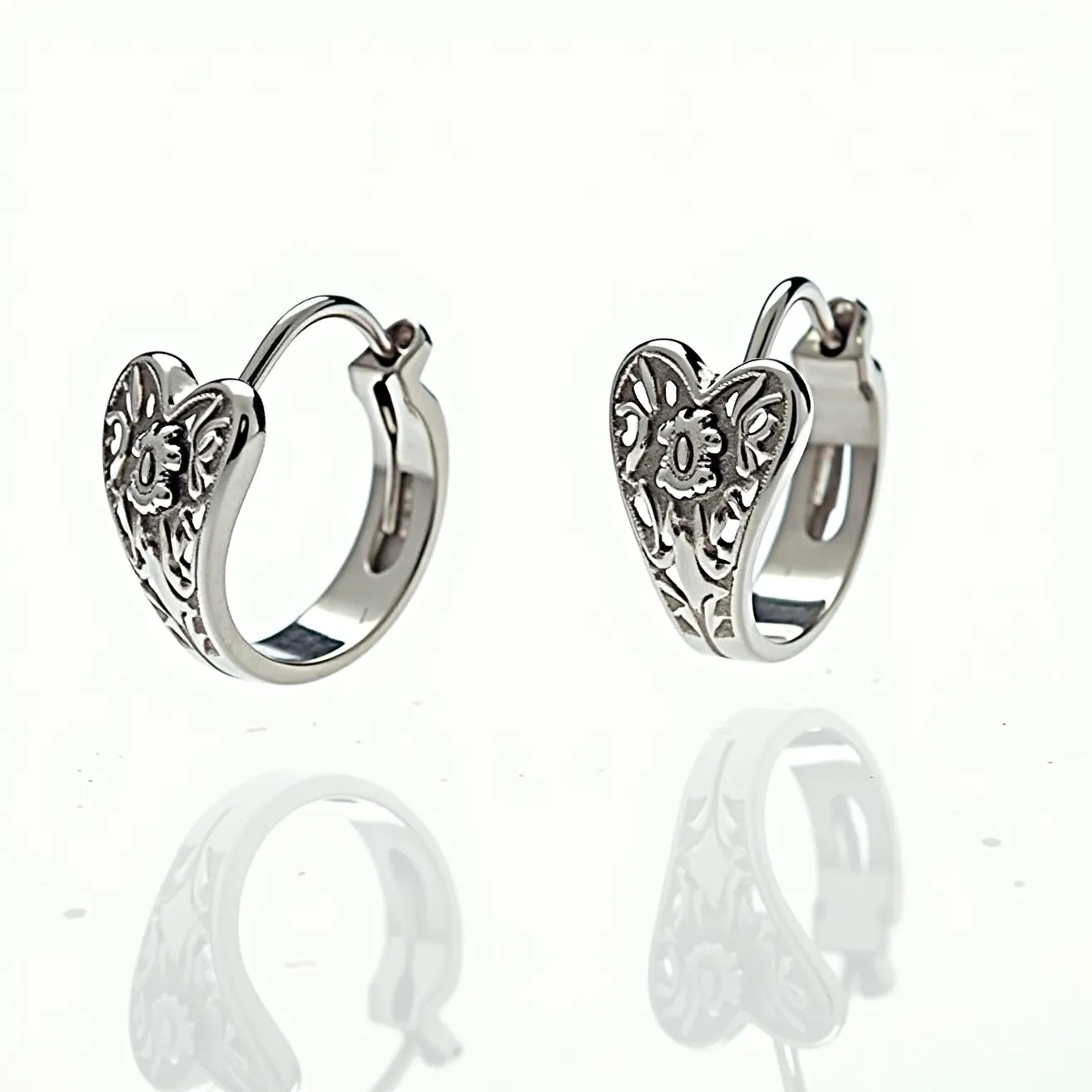 These heart hoop earrings are crafted from a polished metallic material, likely silver or white gold, giving them a shiny and elegant appearance. The design features an engraved heart pattern with intricate floral motifs, providing a classic and ornate aesthetic. There are no visible gemstones incorporated into the design, emphasizing the metalwork's artistry. The earrings utilize a hinge clasp mechanism, allowing for secure and comfortable fastening on the earlobe.