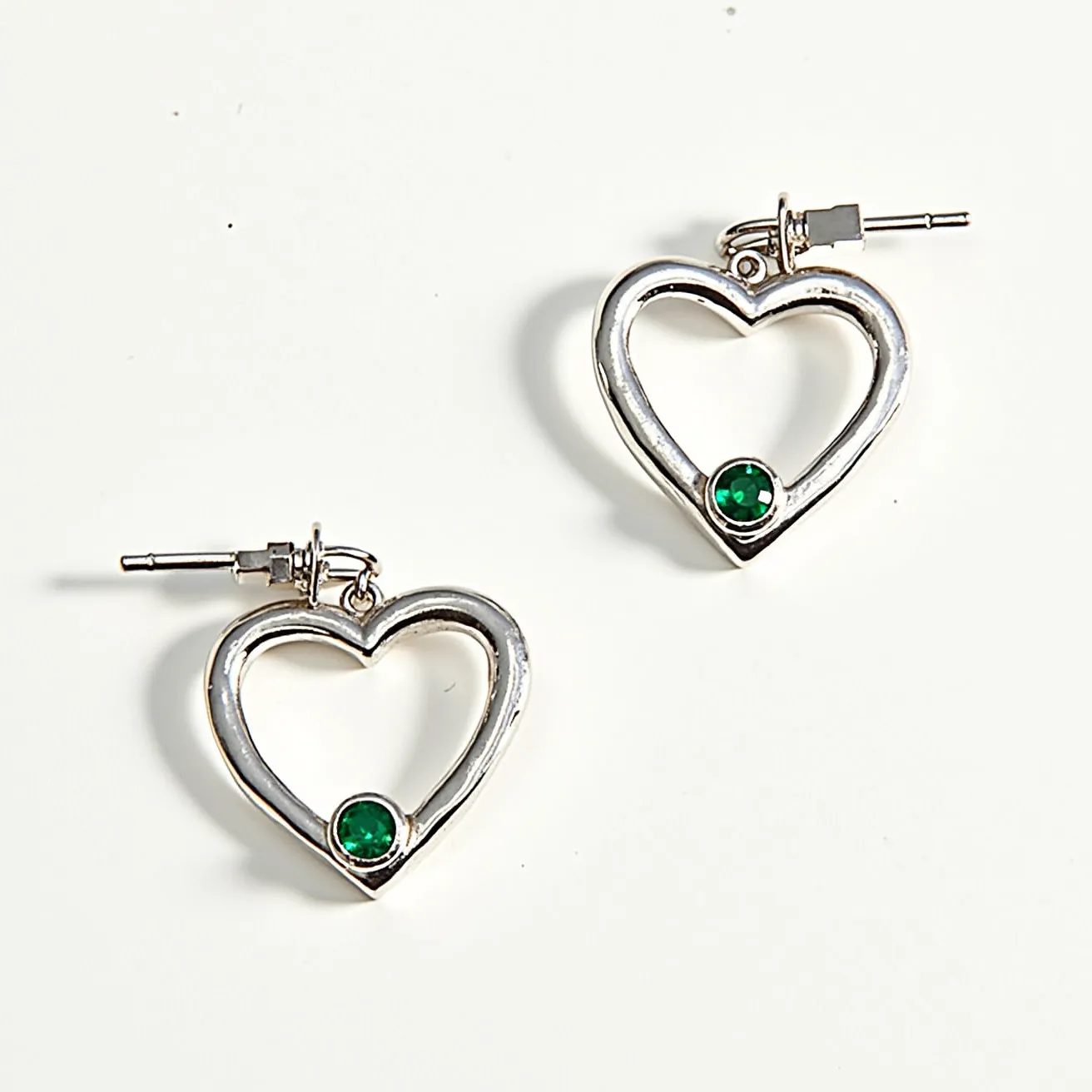 These heart hoop earrings feature a polished silver-tone metal forming the shape of a heart. At the base of each hoop, a round green gem is set, likely an emerald, secured in a bezel setting that complements the overall design. The earrings utilize a post and hinge closure, providing a secure attachment. The sleek, open design of the heart hoops allows the gemstones to stand out as a central feature, combining elegance with a touch of color.