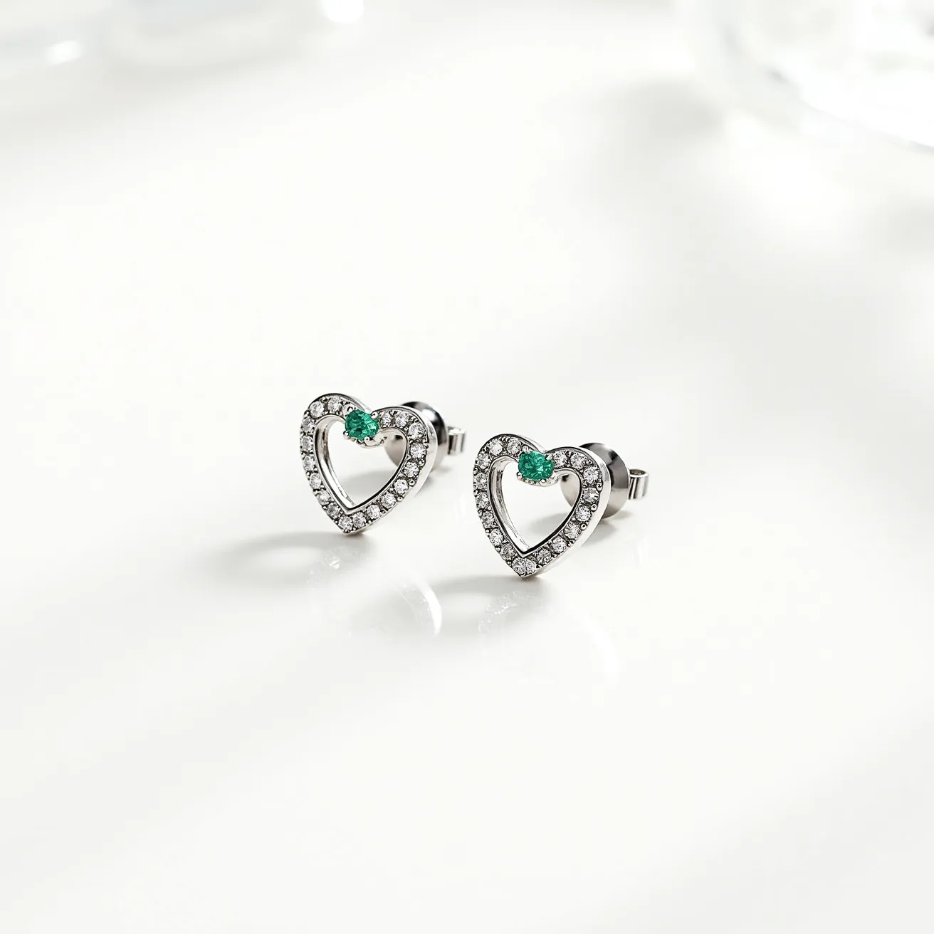 These heart stud earrings feature a delicate heart-shaped design crafted from a polished metal, likely silver or white gold, providing a sleek and shiny finish. The outline of each heart is encrusted with small, sparkling clear stones, possibly diamonds or cubic zirconia, set in a pave style to maximize brilliance and coverage. At the top corner of each heart sits a singular vivid green gem, possibly an emerald, cut in a round or similar shape, secured with a prong setting to highlight its vibrant hue. The earrings are attached via a standard post, complete with a butterfly or friction back clasp, ensuring a secure and comfortable fit.