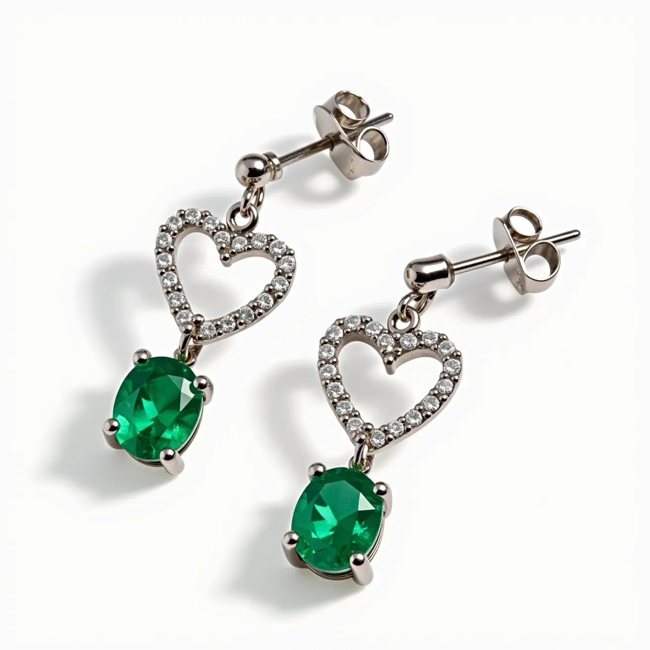 These heart stud earrings feature open-heart shapes encrusted with small, round-cut clear gemstones, set in a metal that appears silvery and reflective, suggesting it could be a type of white gold or sterling silver. Below each heart hangs a vivid green, oval-cut gemstone secured by four prongs, enhancing its vibrant color. The earrings have a classic post and butterfly backing, ensuring a secure yet comfortable fit when worn. The overall design combines elegance with a touch of modern charm, making them suitable for both casual and formal occasions.