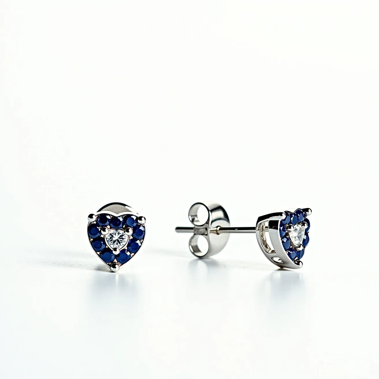 These heart stud earrings feature a central clear gemstone, possibly a diamond, with a brilliant cut that reflects light beautifully. The surrounding halo consists of deep blue stones, likely sapphires, arranged in a heart shape that enhances the central gem's appeal. The setting is crafted from a lustrous metal, possibly white gold or platinum, which complements the stones' colors. The attachment is a classic post with a butterfly back, ensuring a secure fit for the wearer.