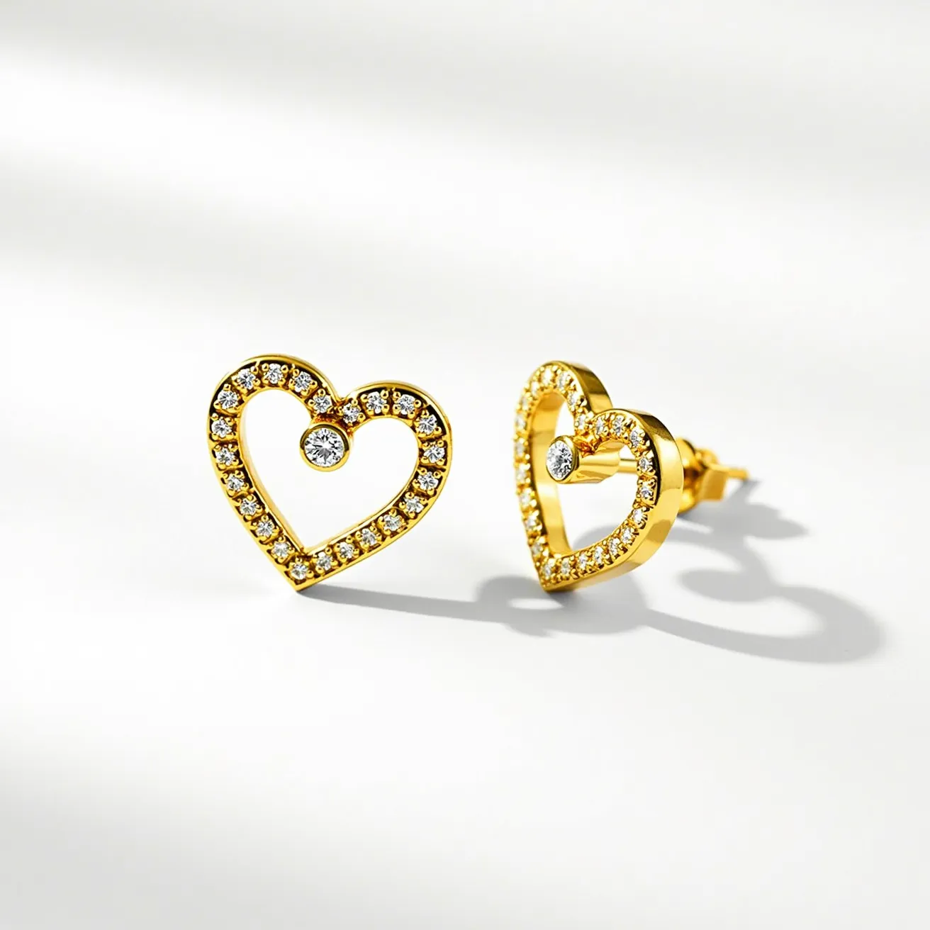 These heart stud earrings feature a delicate gold metal framework forming an open heart shape. Each earring is accentuated with numerous small, round-cut diamonds along the perimeter, offering a sparkling effect. In the center of each heart, a larger diamond is set in a classic prong setting, adding a focal point that draws attention. The earrings utilize a standard post and butterfly clutch back for secure attachment. The combination of gold and diamonds creates an elegant and timeless appeal suitable for various occasions.