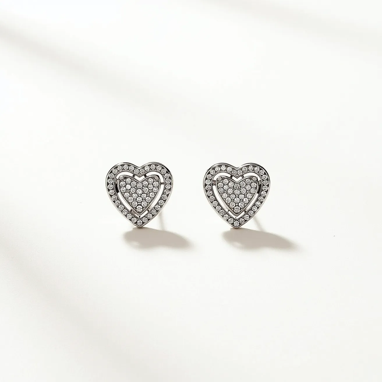 These heart stud earrings feature a sophisticated design with a metal base that appears silvery, possibly white gold or platinum. The earrings are embellished with numerous small, round-cut gemstones, likely diamonds or cubic zirconia, set pave-style across the heart shape, creating a dazzling effect. The studs have a symmetrical, double-heart structure, with a smaller heart encompassed by a larger one, both adorned with gemstones. The attachment is a traditional post, suitable for pierced ears, likely secured with a push back or butterfly clasp, ensuring a secure fit on the earlobe while maintaining elegance and sparkle.