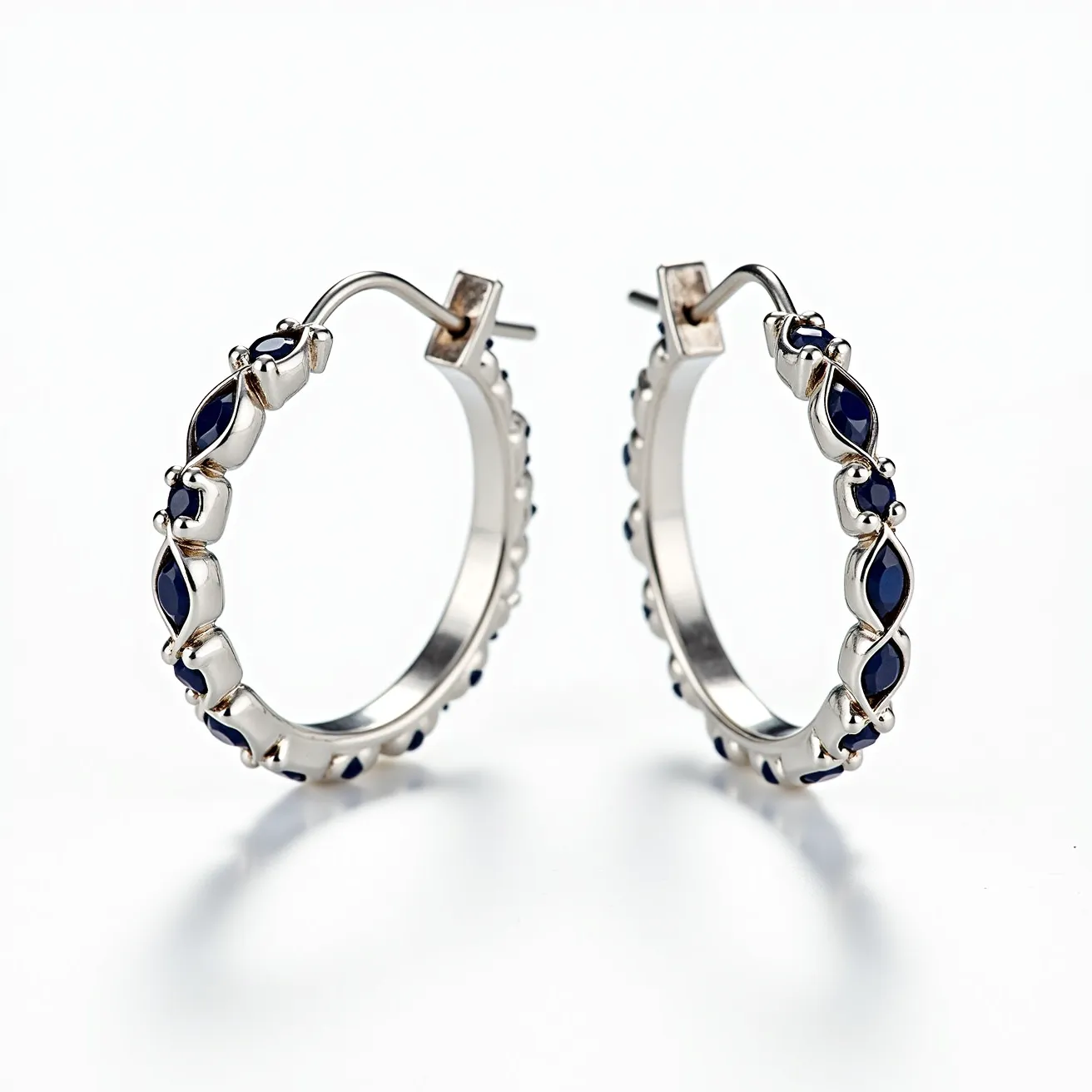 These helix hoop earrings feature a sleek metal construction, likely silver or white gold, forming a circular design that embraces elegance. Along the outer edge of each hoop is a series of deep blue marquise-cut stones, possibly sapphires, set individually in a secure prong setting that showcases their rich hue. The earrings are designed with a hinged post and latch back clasp, providing stability and ease of wear, ensuring they sit securely in the ear. The intricate interplay of metal and gemstones adds a touch of sophistication and allure to the overall design.