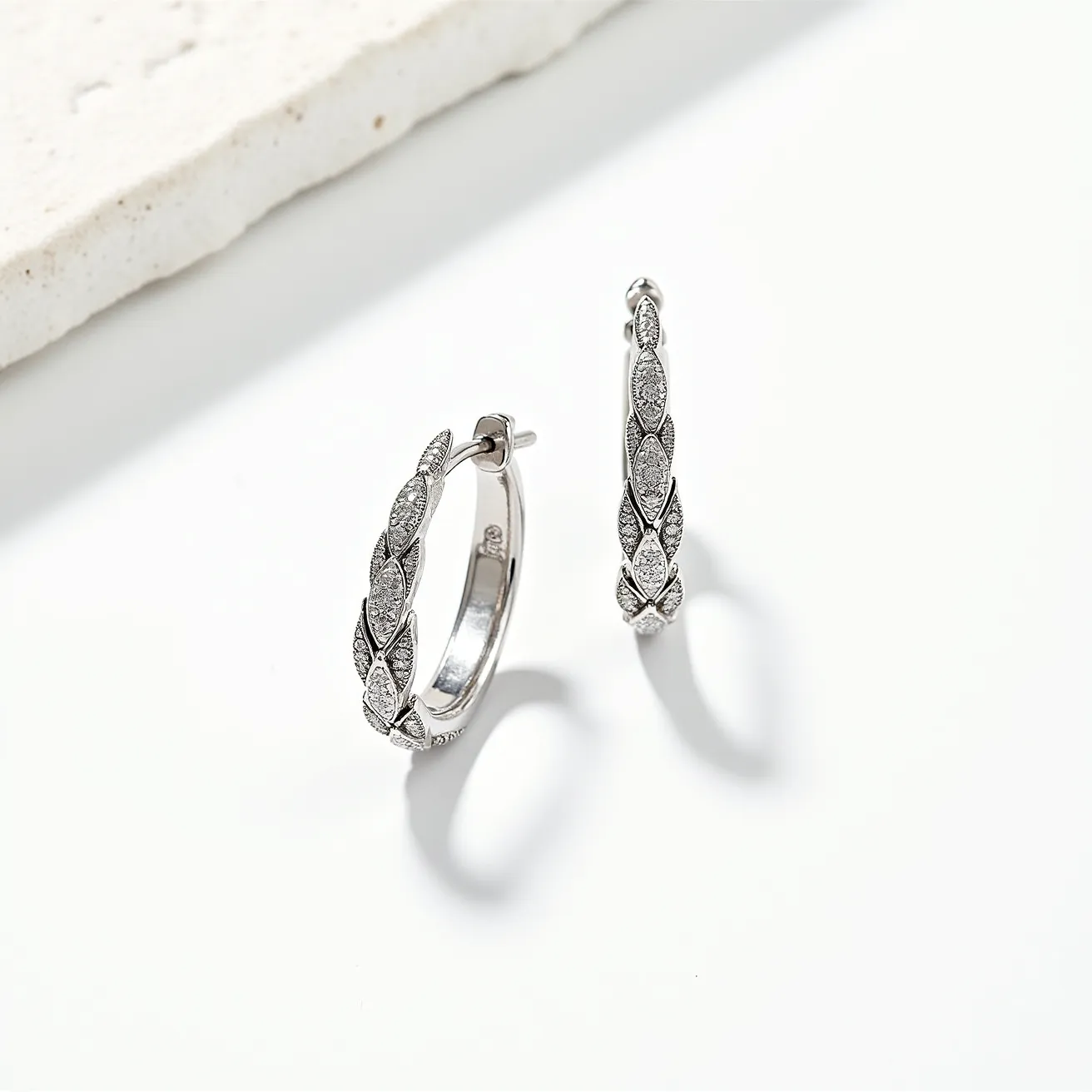 These helix hoop earrings are crafted from a shiny metallic material, likely silver or white gold, providing a sleek and polished look. The design features an intricate pattern of leaf-like shapes that smoothly spiral around the band, encrusted with small, sparkling stones, possibly diamonds, set in a pave style that adds to the overall luxurious appearance. The earrings utilize a post and latch-back closure, ensuring secure attachment while being comfortable to wear. The combination of the textured detailing and the glimmering stones creates an elegant and sophisticated aesthetic.