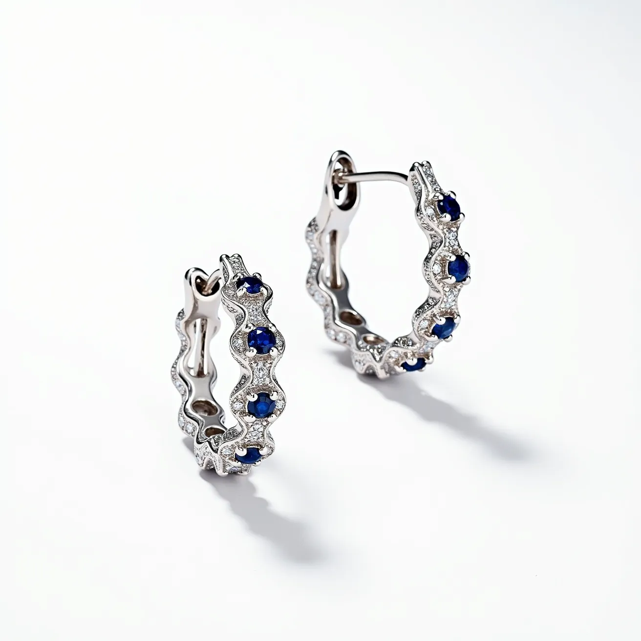 These helix hoop earrings feature a striking design crafted from polished metallic material, likely white gold or sterling silver, that forms a wavy, helix shape. The earrings are elegantly adorned with blue gemstones, possibly sapphires, showcasing a round cut that captures light and adds a vibrant splash of color. These stones are nestled within a pave setting, which is embellished with smaller clear stones, potentially diamonds or cubic zirconia, providing a shimmering contrast. The earrings are secured with a hinged clasp mechanism, ensuring a snug and stylish fit for the wearer.