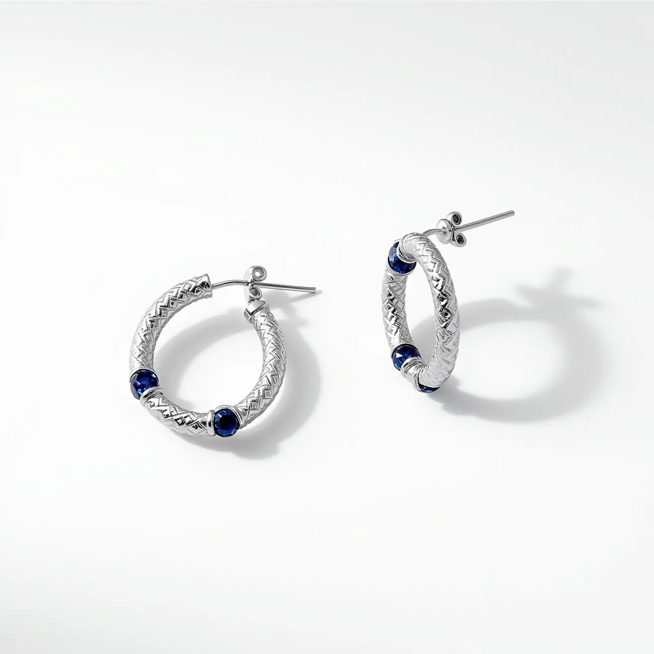 These helix hoop earrings are crafted from a shiny metallic material, likely silver or white gold, featuring a textured, braided design that adds depth and elegance. Each hoop is accented with multiple deep blue gemstones, possibly sapphires, set into the earrings in a bezel setting that securely holds each stone while allowing their facets to catch the light. The hoops are designed for pierced ears, utilizing a straight post and back clasp attachment for secure and comfortable wear. The combination of the textured metal with the rich blue stones creates a sophisticated and modern aesthetic.