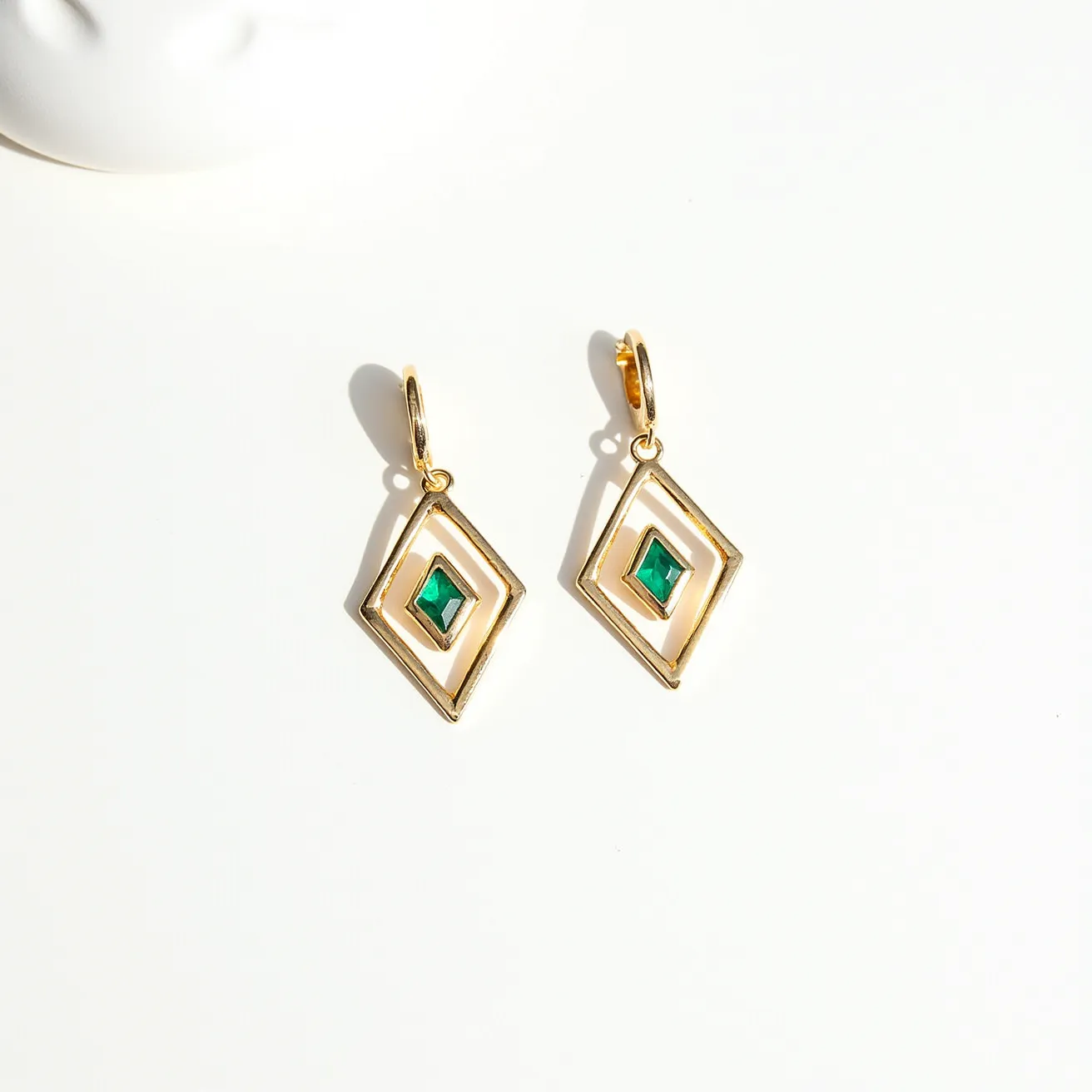 These hoop earrings feature a geometric design with a gold-toned metal framework, shaped into elongated diamond forms. At the center of each hoop is a bright green square-cut stone, securely set within the inner open section of the earrings, adding a pop of color. The earrings are equipped with a simple hoop clasp mechanism for easy attachment and secure wear. The interplay of the gold framework and vibrant stones creates an elegant and modern aesthetic.