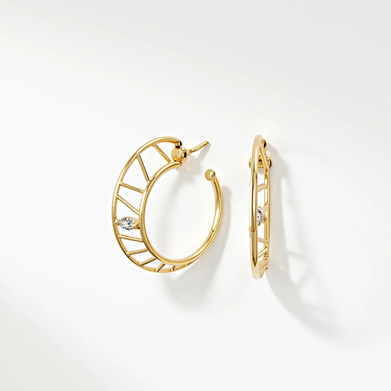 These hoop earrings are crafted from a polished gold-toned metal, featuring an open lattice design. They are adorned with a single marquise-cut gem set into each earring, adding a touch of sparkle. The clasp is a simple post-back that secures comfortably and seamlessly. The combination of the open structure and the strategically placed gem creates an elegant and modern aesthetic.