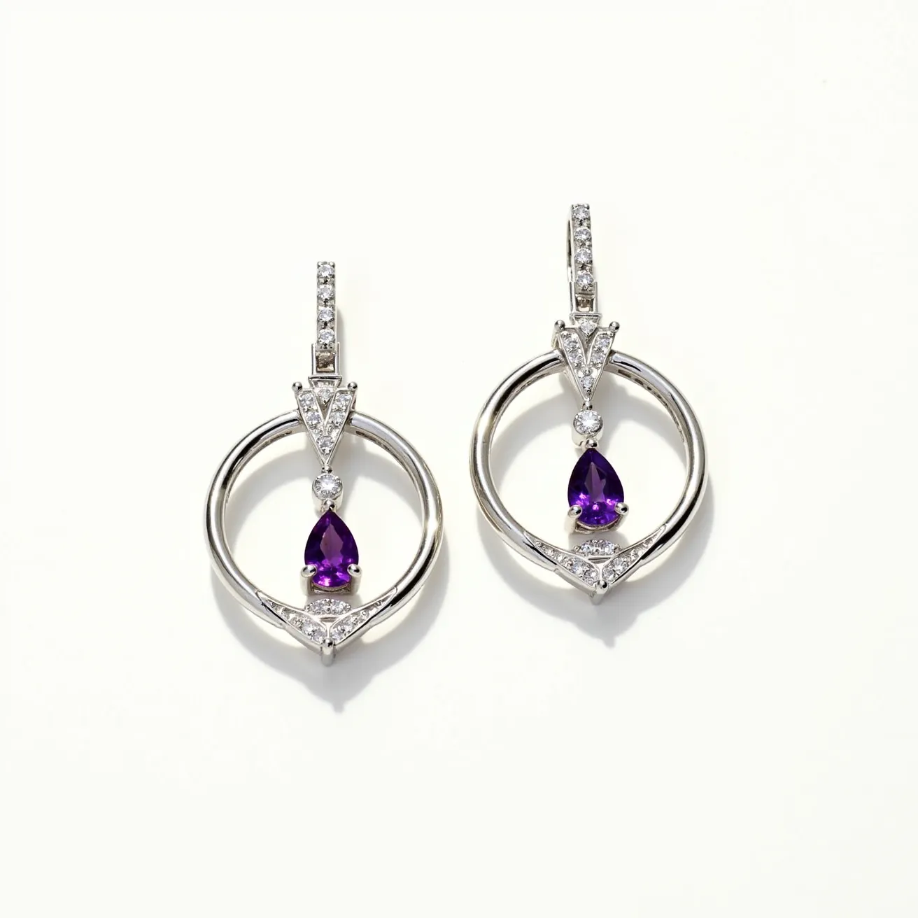 These hoop earrings feature a sleek circular design crafted from polished metal, likely silver or white gold, giving them a refined and elegant look. Within the hoop, a striking pear-shaped purple gemstone, possibly an amethyst, is set prominently, drawing attention with its deep hue. The gemstone is suspended within the hoop and secured by a prong setting that adds to its brilliance. Accentuating the design are small clear stones, most likely diamonds, arranged in a triangular cut above the central gemstone and along the ear clasp, adding sparkle and sophistication. The earrings are fitted with a hinged lever-back clasp, ensuring a secure and comfortable fit when worn.