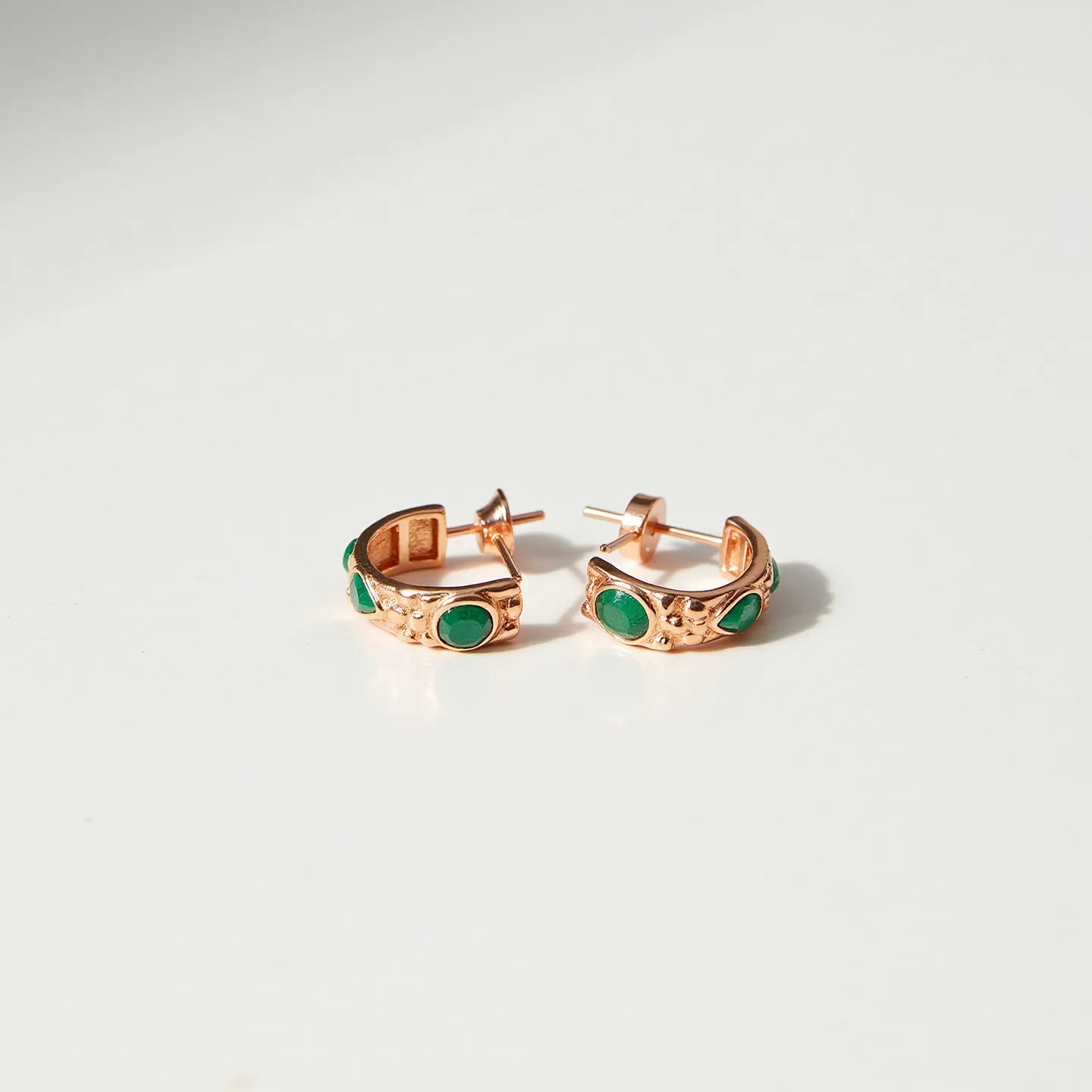 These hoop earrings are crafted from a warm, rose gold material, featuring vibrant green stones set into the metal. The stones, with a round cut, are bezel set, adding a pop of color to the elegant design. The earrings are secured with a classic post and friction back clasp, ensuring a secure fit when worn. The detailing on the hoops adds texture and enhances the overall aesthetic appeal, making them a stylish accessory.