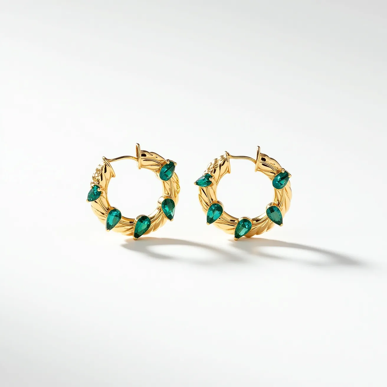 These hoop earrings are crafted from a gold-toned metal, featuring a textured design that adds elegance to the piece. The earrings are adorned with pear-shaped green gemstones, each set securely into the gold material. These stones are evenly spaced around the hoop, enhancing the luxurious appearance. The earrings use a classic latch backing to secure them in place when worn, ensuring both style and functionality.