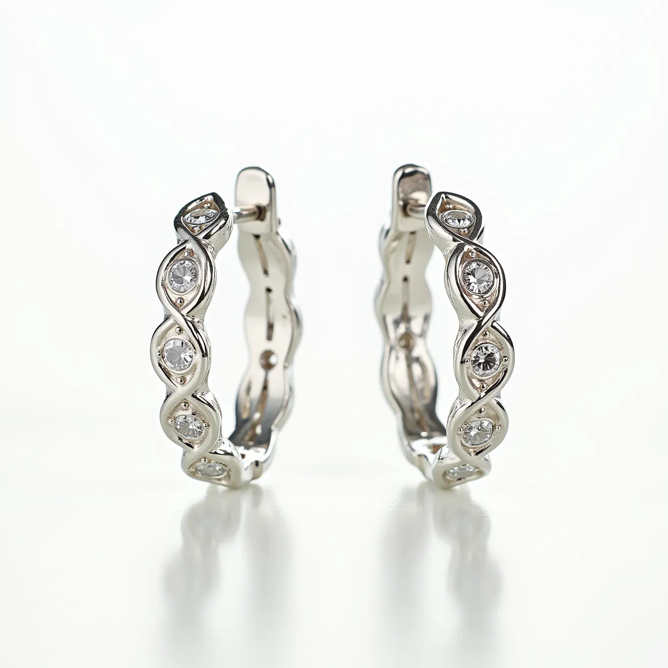 These hoop earrings feature a sophisticated design crafted from what appears to be a polished metal, likely silver or white gold. Embedded in a twisted, elegant pattern are several round-cut gems that resemble diamonds, each set in a secure bezel setting that enhances their brilliance. The attachment consists of a hinge clasp, offering a seamless and secure fastening method. The overall style is both contemporary and classic, making the earrings versatile for various occasions.