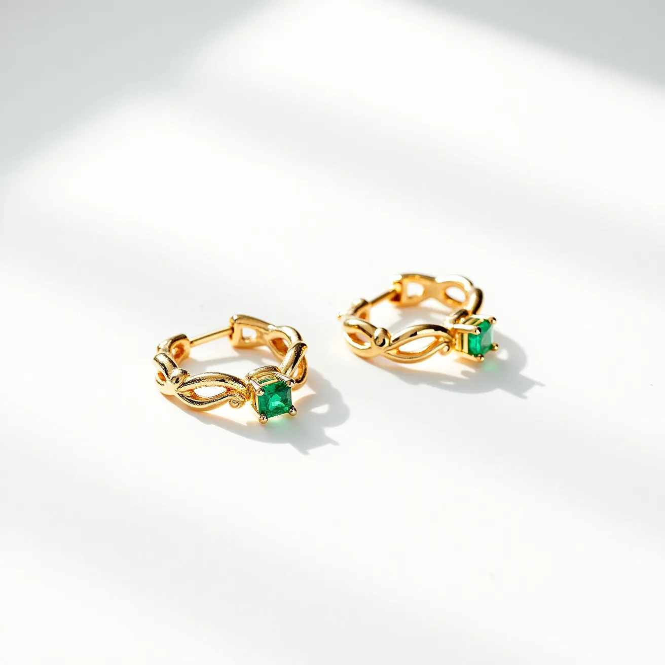 These hoop earrings are crafted from gold-toned metal, featuring an intricate design that interlaces throughout the circular shape. They are adorned with a prominent green stone set in the center, likely an emerald or emerald-like gem, presented in a secure prong setting to enhance its brilliance. The earrings use a hinged clasp for secure attachment, blending functionality with the aesthetic appeal of the ornate design. The overall look is sophisticated, giving these earrings a luxurious feel suitable for elegant occasions.