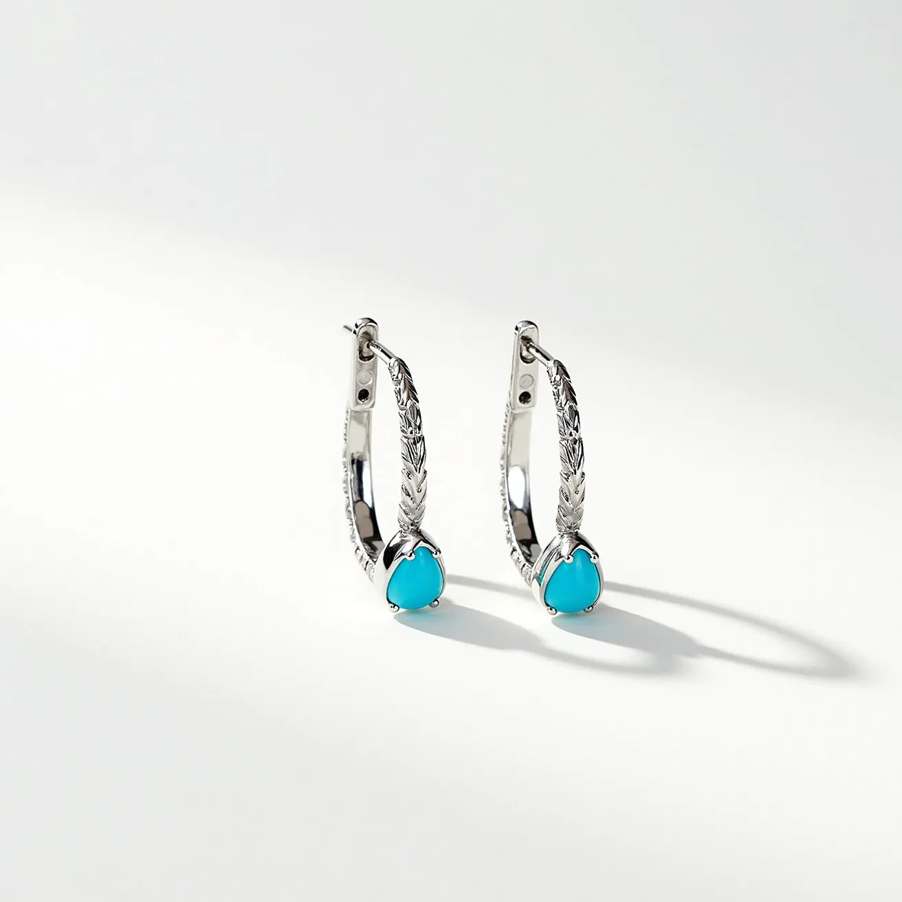 These hoop earrings feature a silver-toned metal construction with an intricate embossed design along the outer surface, enhancing their elegance. Each earring is adorned with a pear-shaped turquoise stone set in a bezel setting, adding a striking contrast to the silver base. The earrings employ a hinge clasp mechanism for secure attachment. The combination of the vibrant turquoise stones and the detailed silver work gives these earrings a distinctive and sophisticated appearance, suitable for both contemporary and traditional styles.