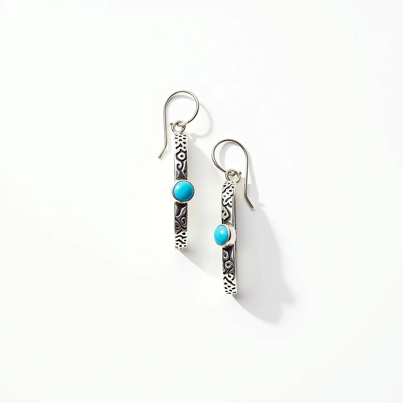 These hoop earrings feature an elongated design crafted from silver with intricate engraved patterns. They are adorned with round turquoise stones, set centrally within each piece, providing a vibrant contrast to the silver. The stones are secured with a bezel setting, emphasizing their smooth, polished surface. The earrings are equipped with hook clasps, allowing for easy wear and a comfortable fit. The combination of silver and turquoise gives the earrings a stylish and classic look.