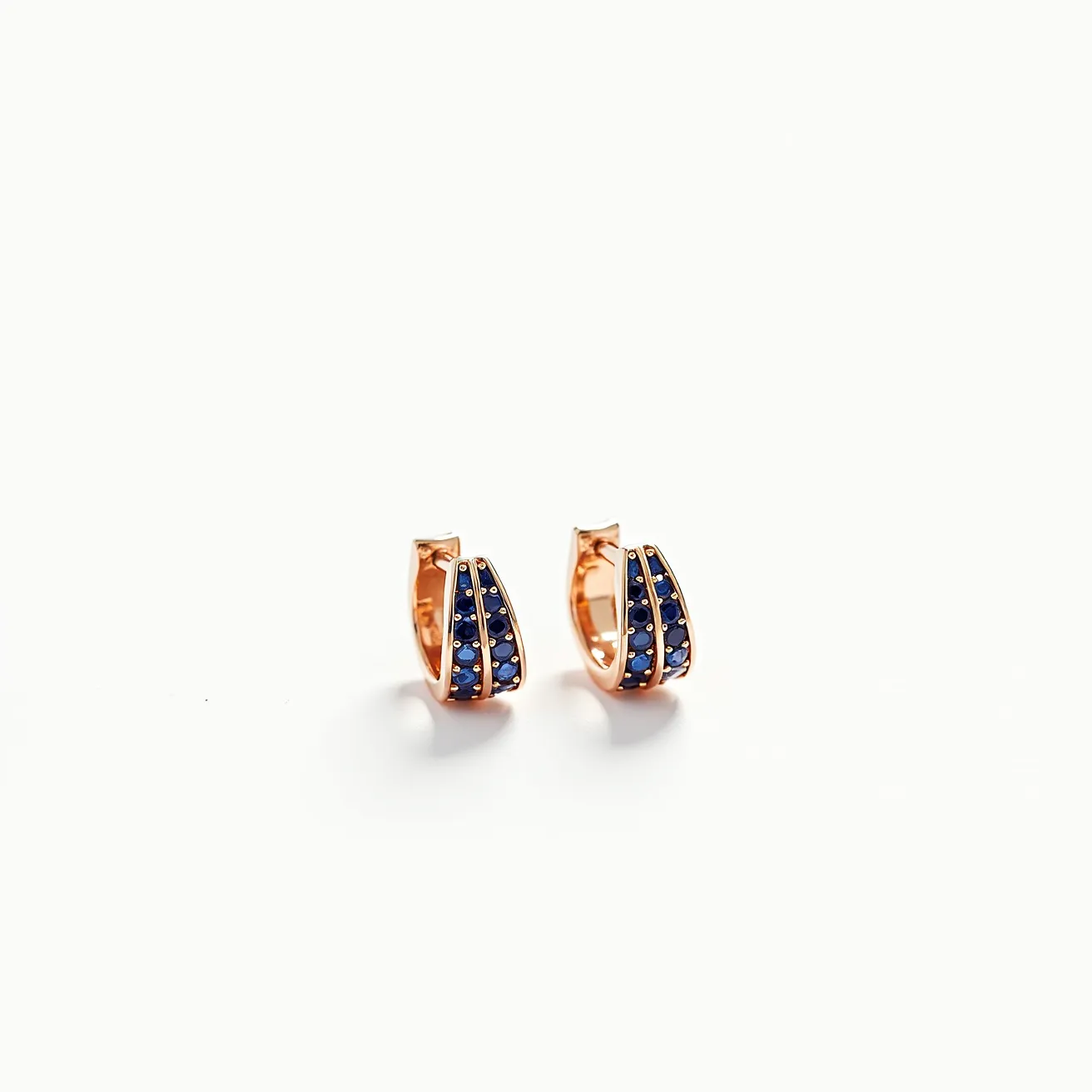 These huggie earrings feature a sleek and elegant design crafted from a warm-toned metal, possibly rose gold or gold-plated material. They are adorned with deep blue gemstones, likely sapphires, arranged in a pavé setting that highlights their vibrant color. The stones are round-cut, adding a classic touch to the modern hoop style. These earrings utilize a hinge clasp mechanism, ensuring secure and comfortable wear. The thoughtful arrangement of the gems creates an appealing visual contrast against the metallic base, making these earrings a striking accessory.