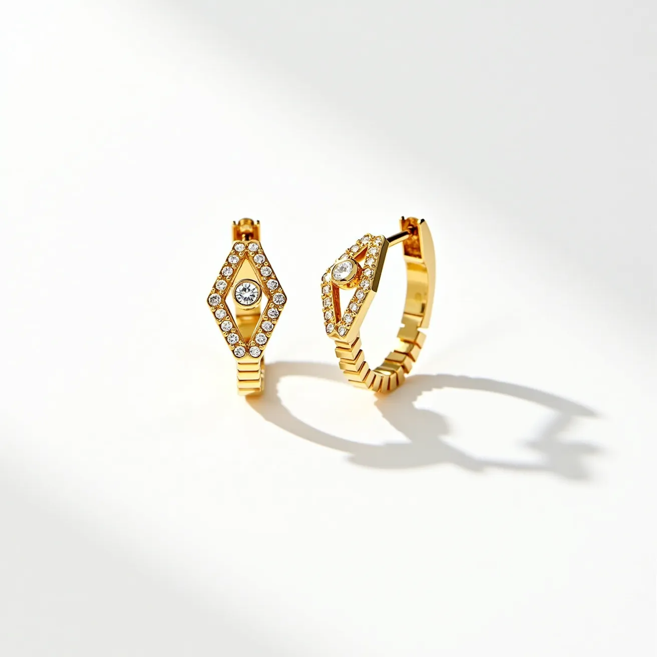 These huggie earrings are crafted from gold, showcasing a sleek, modern design. Each earring is adorned with a prominent central round-cut diamond held securely in a bezel setting, surrounded by smaller pave-set diamonds that add extra sparkle. The earrings feature a hinged clasp mechanism that ensures a secure and comfortable fit around the earlobe. The overall look is elegant and sophisticated, highlighting a geometric design with its diamond-shaped arrangement.