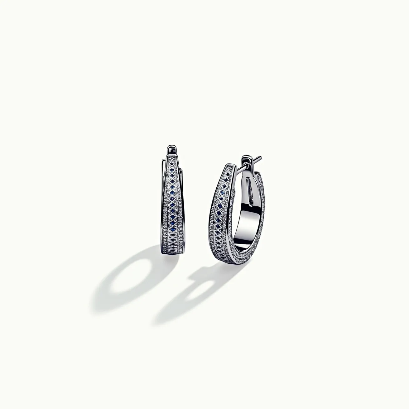 These huggie earrings feature an elegant design crafted from a shiny metal, likely white gold or platinum, which provides a sophisticated backdrop for the embedded gemstones. The earrings are adorned with a series of round-cut and possibly pavé-set diamonds that shimmer along the outer surface. Additionally, there appear to be small blue stones, such as sapphires, complementing the diamonds and adding a touch of color to the design. The earrings are designed with a secure hinged clasp mechanism that ensures ease of wear while maintaining a sleek, continuous hoop appearance. These elements together create a luxurious and timeless accessory.