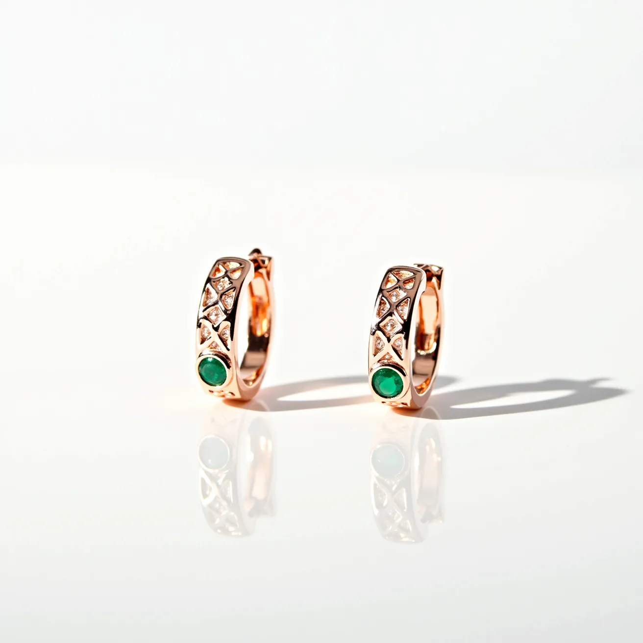 These huggie earrings are crafted from a warm-toned metal, possibly rose gold, giving them an elegant and luxurious appearance. The design features a vibrant green gemstone at the base of each hoop, set in a bezel setting that secures the stone while showcasing its round cut. Above the green gemstone, intricate diamond-shaped accents are inlaid with smaller, clear stones, likely diamonds, which are set in a pavé style to add sparkle and texture. The earrings are designed with a hinged clasp, allowing for easy and secure attachment to the ear lobe, ensuring both comfort and style.