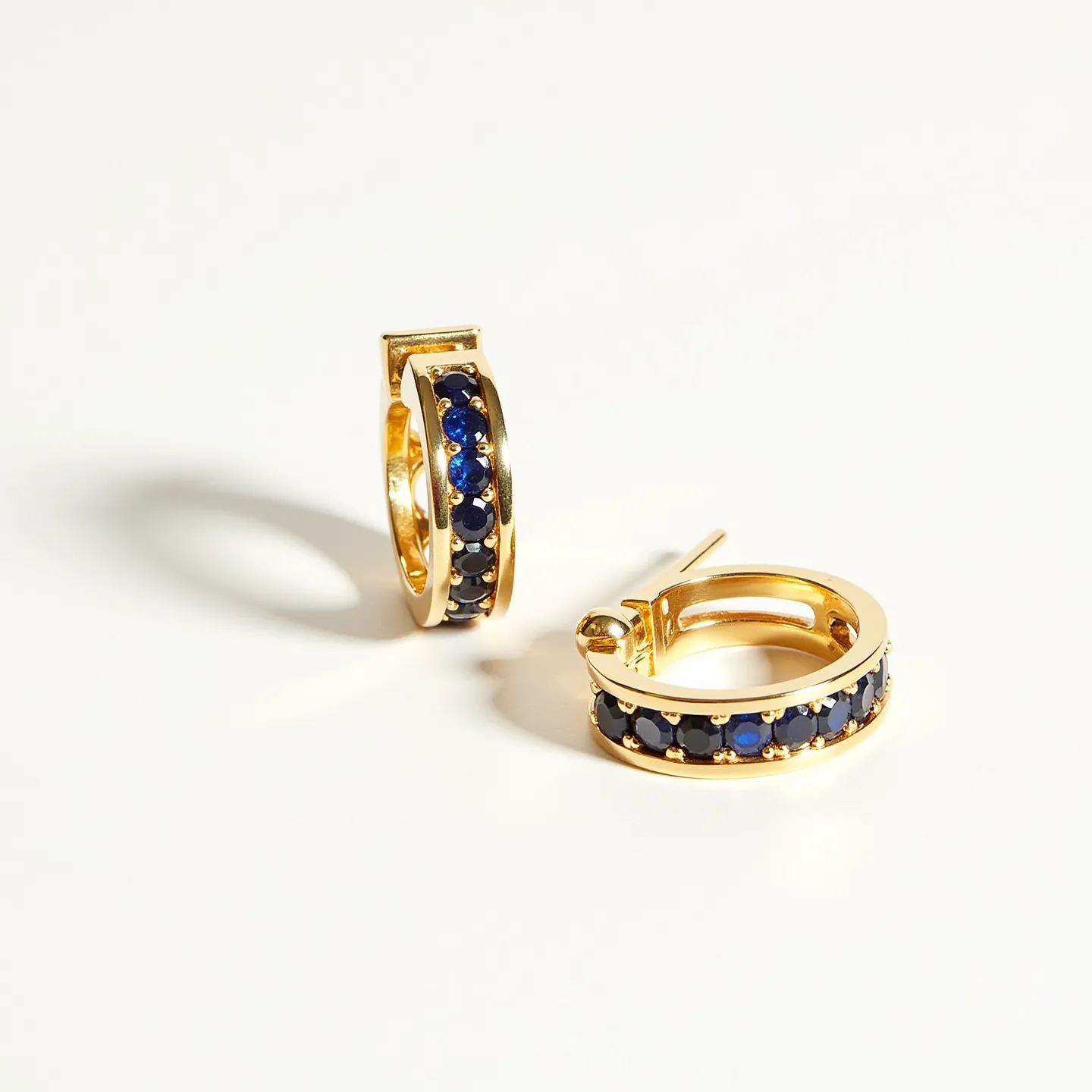 These huggie earrings feature a sleek and elegant design, crafted from a gold-toned metal. The hoops are adorned with a series of round, deep blue gemstones, likely sapphires, each meticulously set in a shared prong setting that enhances their sparkle. The stones are closely set along the outer curve of the earrings, adding a touch of opulence. The earrings are equipped with a hinge and latch back clasp, providing a secure and comfortable fit for everyday wear.