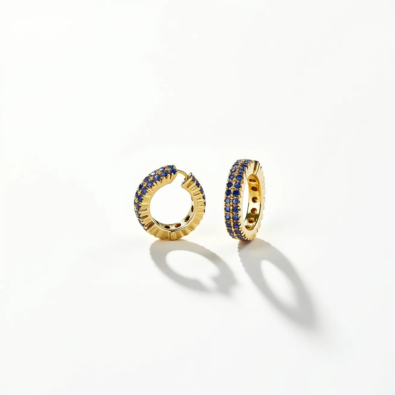 These huggie earrings feature a sleek gold base that elegantly supports rows of small, round blue gemstones, likely sapphires, embedded in a neat pavé setting. The gemstones have a brilliant cut, designed to maximize sparkle and catch the light beautifully. The earrings utilize a hinged clasp mechanism that ensures a secure and comfortable fit on the earlobe, making them both stylish and practical for everyday wear.