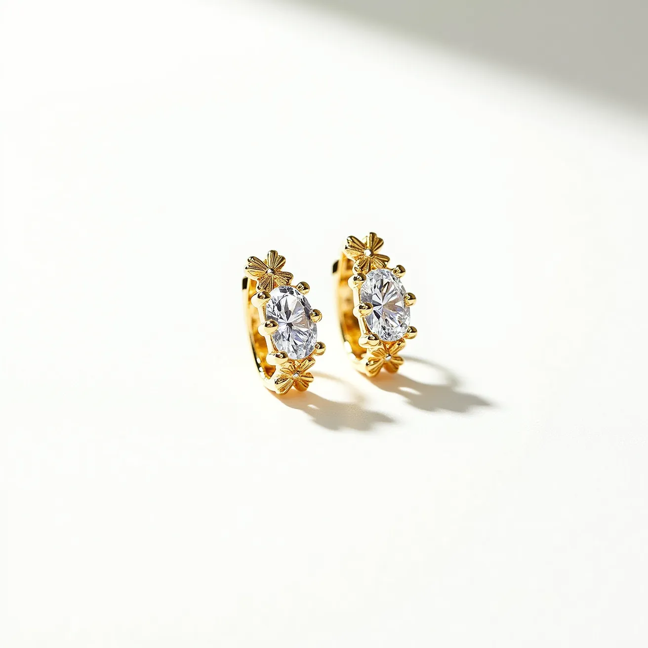 These huggie earrings are crafted from a bright gold-toned metal, featuring an elegant design with a central oval-cut gem that sparkles prominently. Each earring displays a beautiful, clear stone set securely in a prong setting, which enhances its brilliance and holds it firmly in place. Delicate floral motifs decorate the outer edge, adding a touch of sophistication and charm to the overall design. The earrings are designed with a huggie clasp, ensuring a snug and secure fit around the earlobe, making them a stylish and functional accessory.