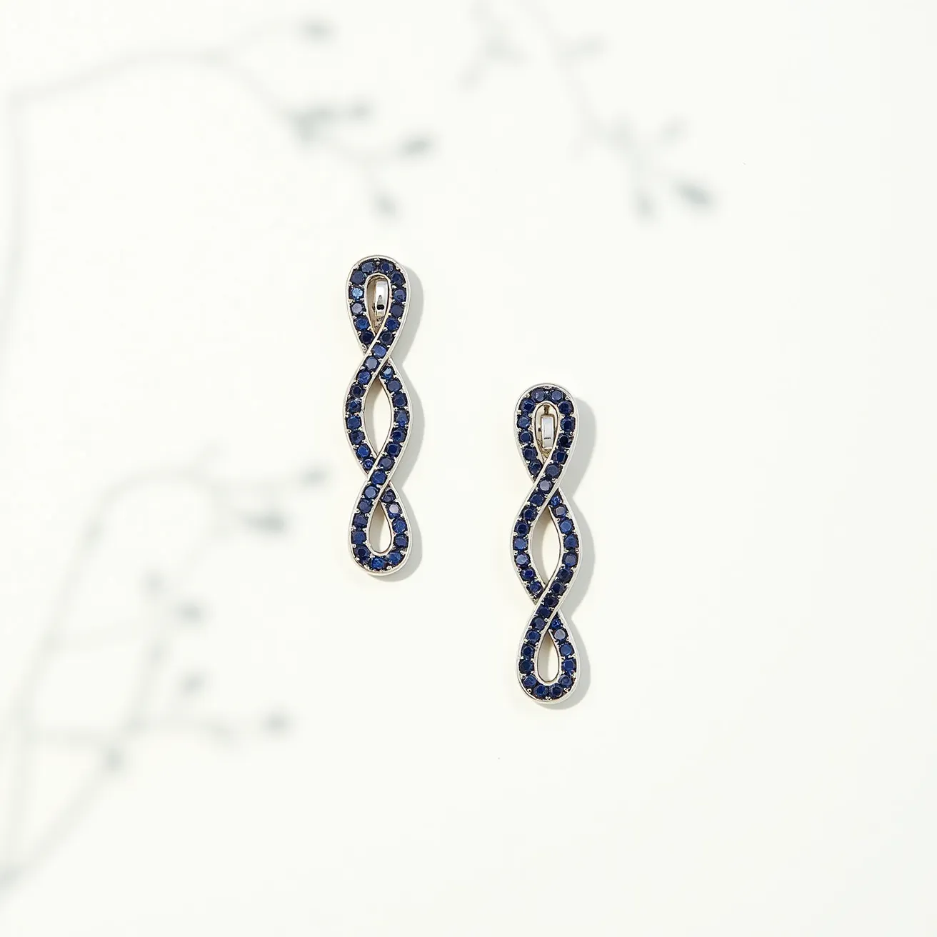 These infinity earrings feature a delicate and elegant design with a continuous, twisted shape forming the infinity symbol. The earrings are adorned with dark blue gems, likely sapphires, which are round-cut and set closely together in a pavé setting. The metal base appears to be a polished silver or white gold, enhancing the brilliance of the stones. These earrings include a simple post for easy attachment, providing a seamless and secure fastening. The overall composition exudes a sophisticated and timeless appeal.
