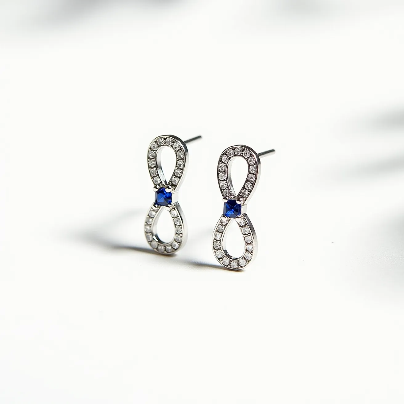 These infinity earrings feature a sleek, silver-colored metal forming the iconic infinity loop design. Each earring is beautifully adorned with small, round, clear stones, likely diamonds or cubic zirconia, meticulously set in a pavé setting throughout the loop. Central to the design is a single, vibrant blue gemstone in a square cut, securely housed in a prong setting, adding a striking contrast to the piece. The earrings attach via simple stud backs, offering a classic and secure fit for elegant wear.