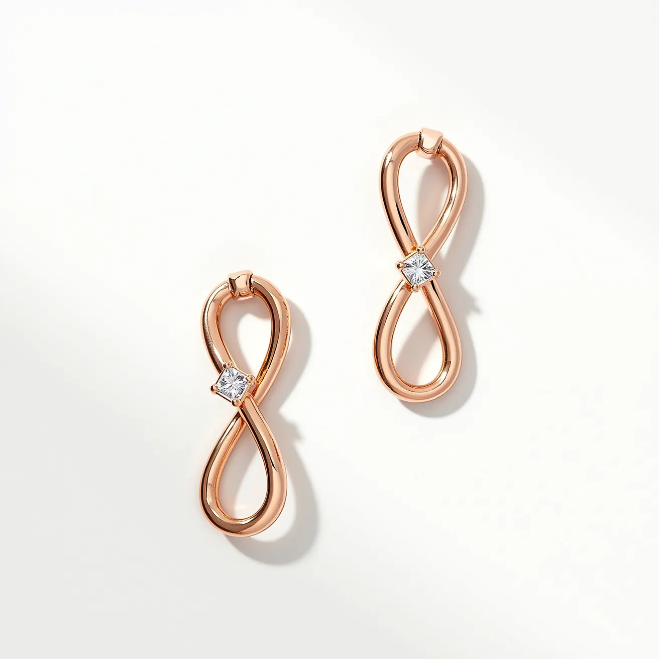 These infinity earrings feature a sleek design crafted from a rose gold material, forming a graceful figure-eight shape. Each earring is adorned with a single, sparkling white gemstone, likely a diamond or cubic zirconia, cut in a square shape and set in a simple prong setting at the center of the infinity loop. The earrings are designed with a post-back attachment, providing secure and comfortable wear. Their elegant and minimalistic style makes them suitable for both everyday wear and special occasions.