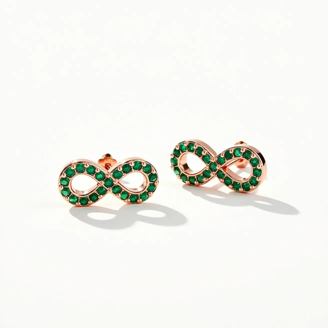 These infinity earrings feature a delicate infinity symbol crafted in a rose gold-tone metal. They are adorned with small, round-cut green gemstones that are evenly set along the loops of the infinity design, adding a vibrant touch to the sleek metal. The earrings are designed with a post-back closure, ensuring a secure and comfortable fit. The combination of the rose gold tone and green gemstones creates a striking contrast, enhancing the elegant and timeless appeal of the earrings.