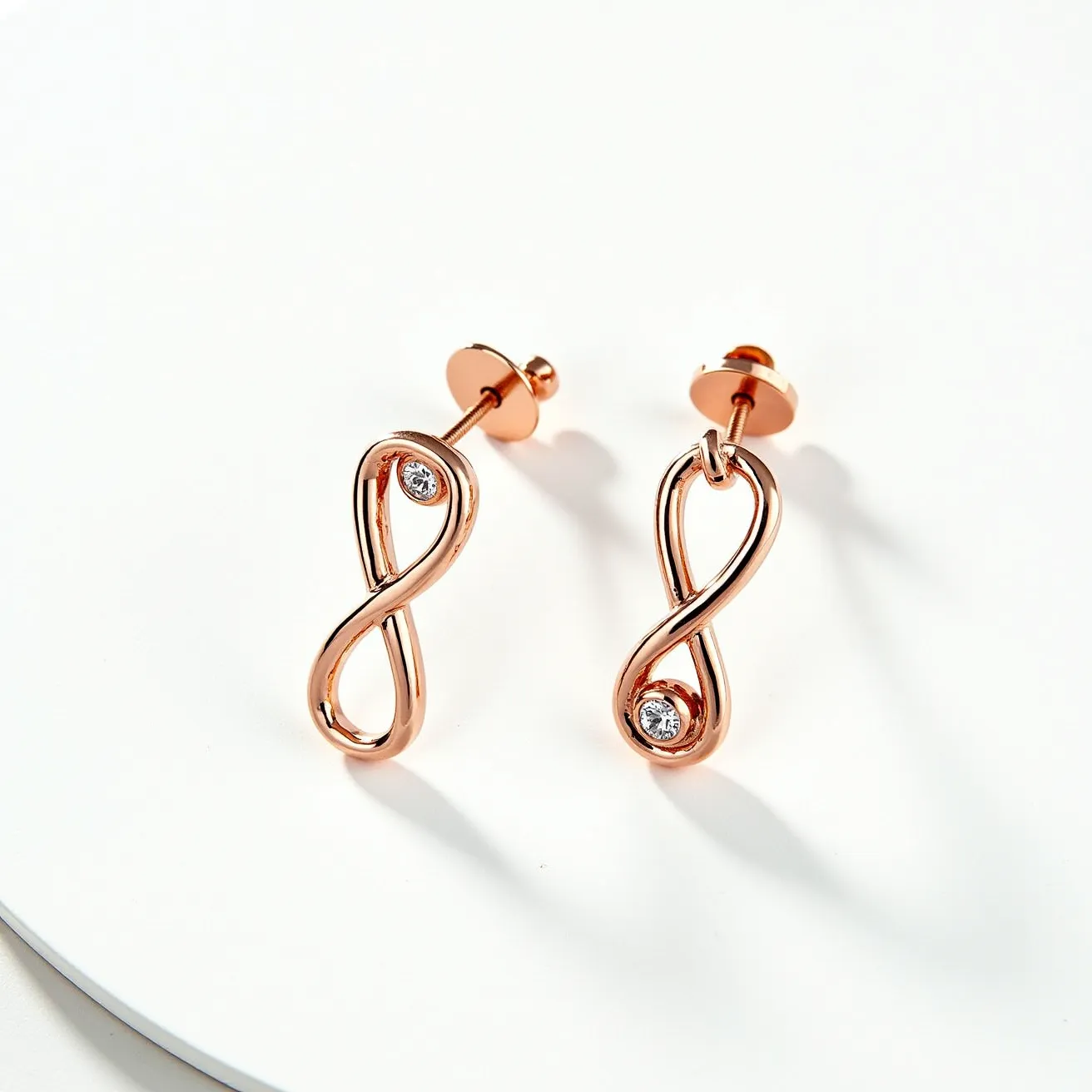 These infinity earrings feature a sleek, rose gold metal, crafted into an elegant infinity symbol. Incorporated within each loop of the symbol is a single, round-cut gem, likely a diamond or a cubic zirconia, set within a bezel setting. The earrings are designed with a secure stud post and a screw-back clasp for attachment, ensuring they stay firmly in place when worn. The polished finish of the metal enhances its reflective quality, complementing the brilliance of the gems.