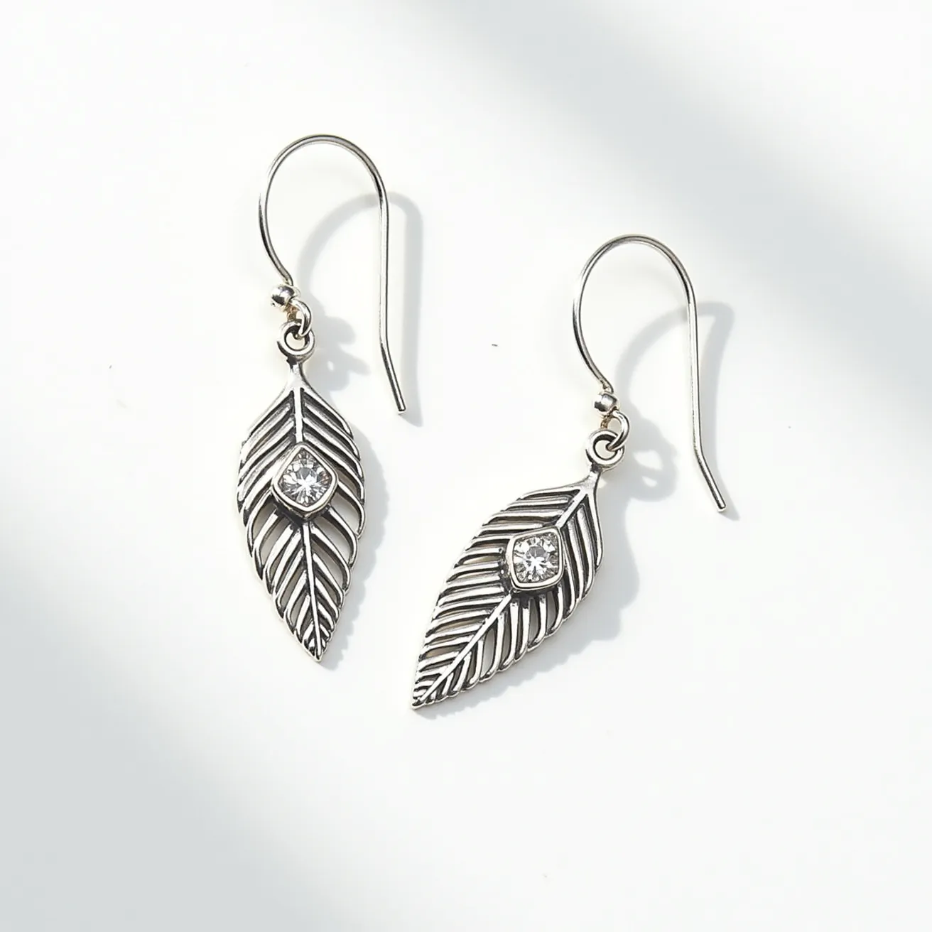 These leaf earrings feature a delicate design with intricately detailed metal resembling the natural veining of a leaf. They are crafted from a shiny, silver-toned metal that adds elegance to their natural motif. At the center of each leaf is a small, round gem, possibly a clear stone like cubic zirconia or a diamond, set in a bezel setting that secures the stone while allowing it to catch the light. The earrings use a simple fishhook clasp for effortless wearability, providing a classic and comfortable fit that dangles elegantly from the ears. The overall craftsmanship highlights both the beauty of nature and sophisticated jewelry design.