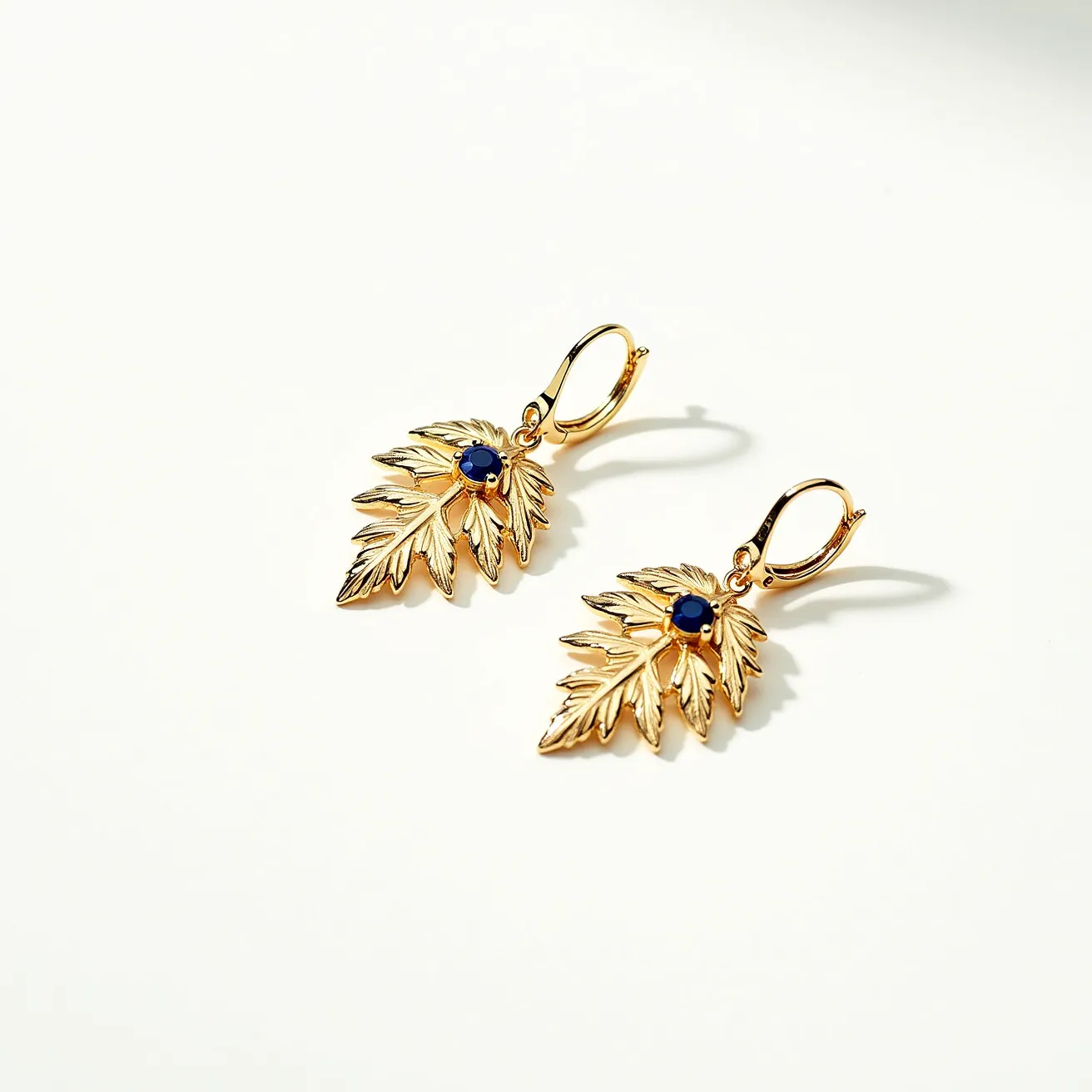 These leaf earrings are crafted from metal with a gold-toned finish, designed to resemble detailed, textured leaves. At the center of each leaf is a small, round, faceted blue gem set in a prong setting, which adds a touch of color and elegance. These earrings feature lever-back clasps, providing a secure and comfortable fit. The intricate design and vibrant gemstone create a striking and elegant accessory suitable for various occasions.