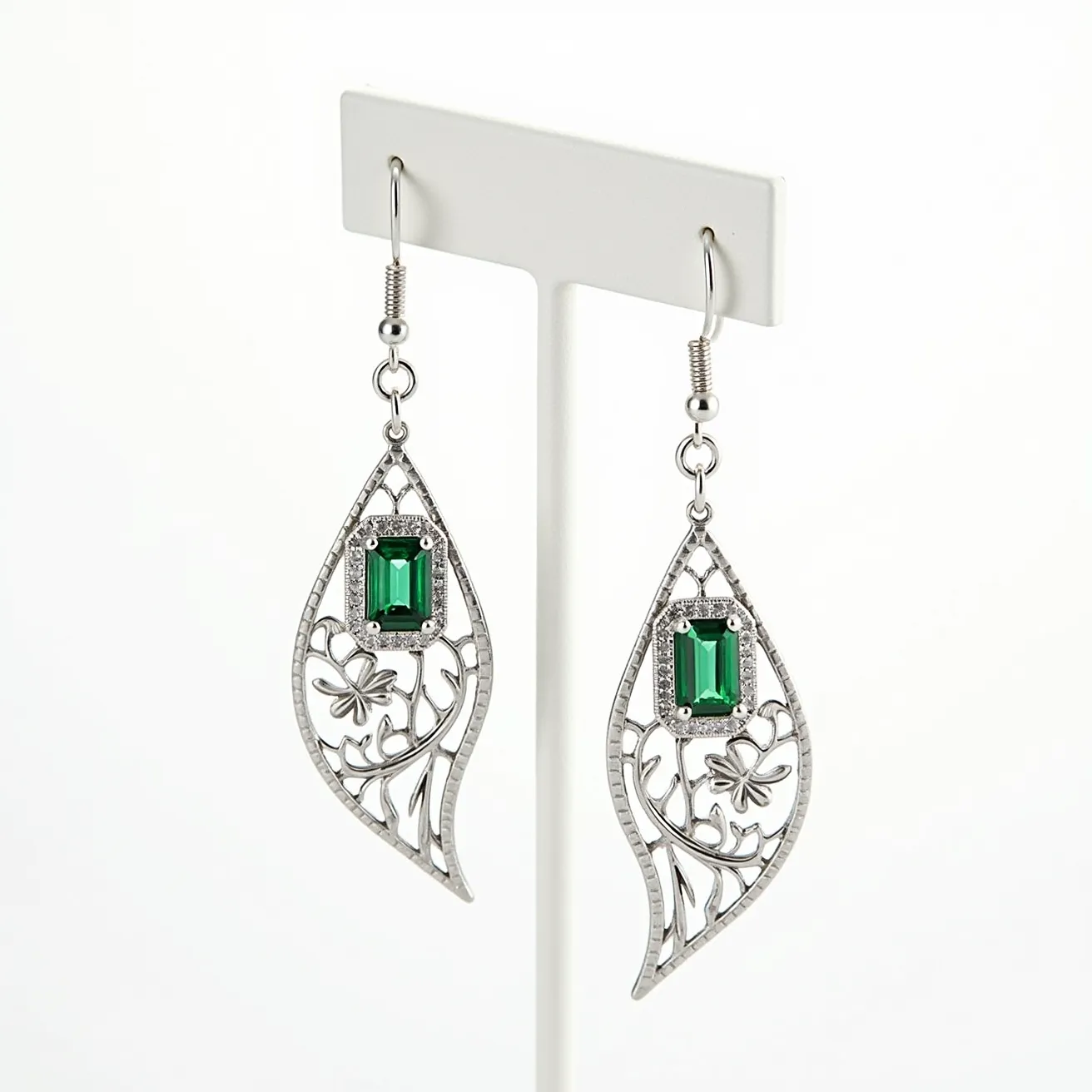 These leaf earrings feature an intricate design crafted in a metallic material, possibly silver, forming an elegant leaf shape. Each earring is adorned with a rectangular emerald-cut green gem set in a bezel, adding a vibrant focal point to the piece. The openwork metal design incorporates delicate floral motifs, enhancing their natural elegance. The earrings are attached with a French hook clasp, providing a secure and comfortable fit.