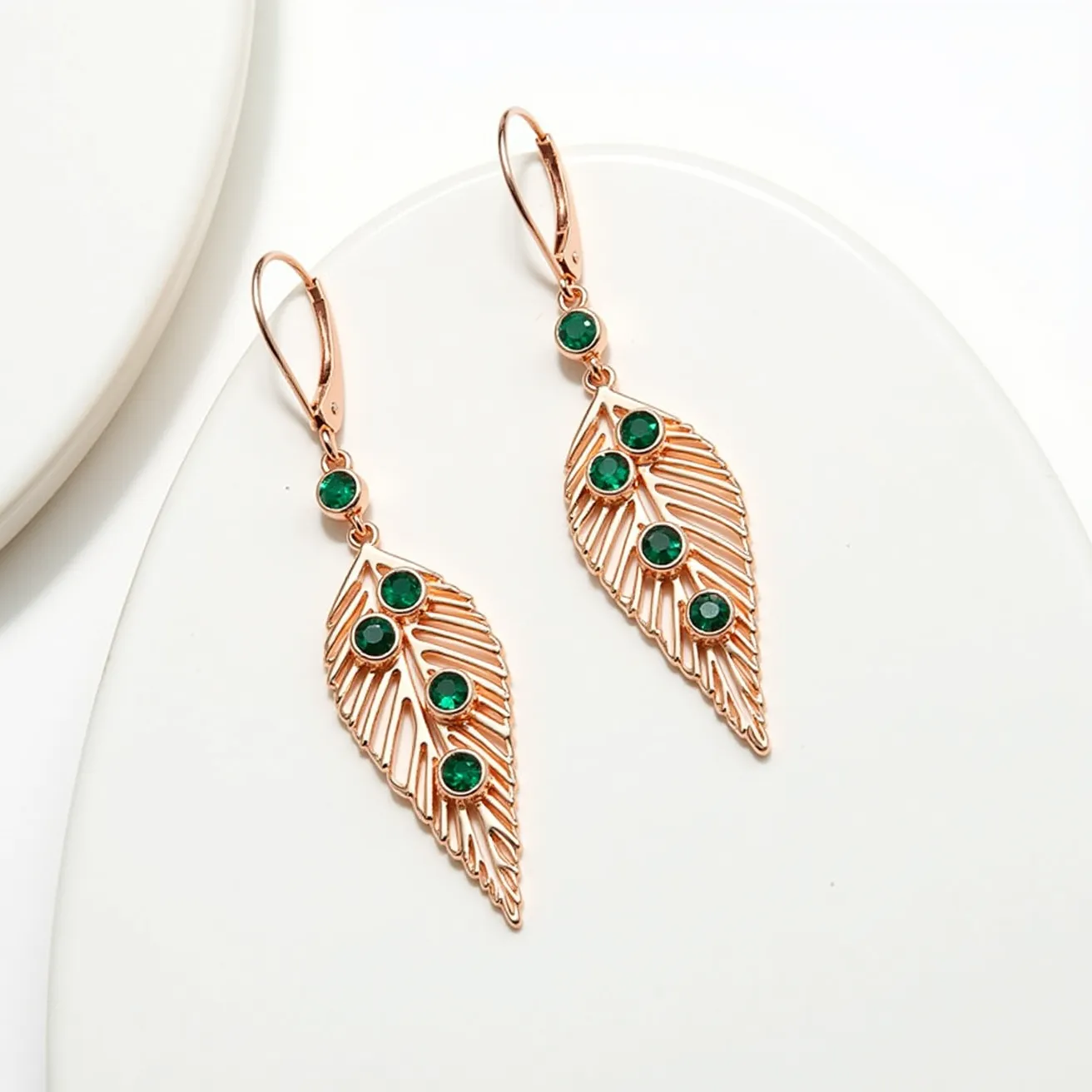 These leaf earrings feature a delicate design with a series of intricate cutouts resembling the veins of a leaf. The material appears to be a warm-toned metal, possibly rose gold or gold-plated, giving them an elegant shine. Adorning the leaves, there are several round, faceted green gems set in bezel settings, adding a touch of color and sparkle to the design. The earrings employ a leverback clasp for secure attachment, ensuring they stay in place while worn. The overall look is both sophisticated and nature-inspired, making them a versatile addition to any jewelry collection.