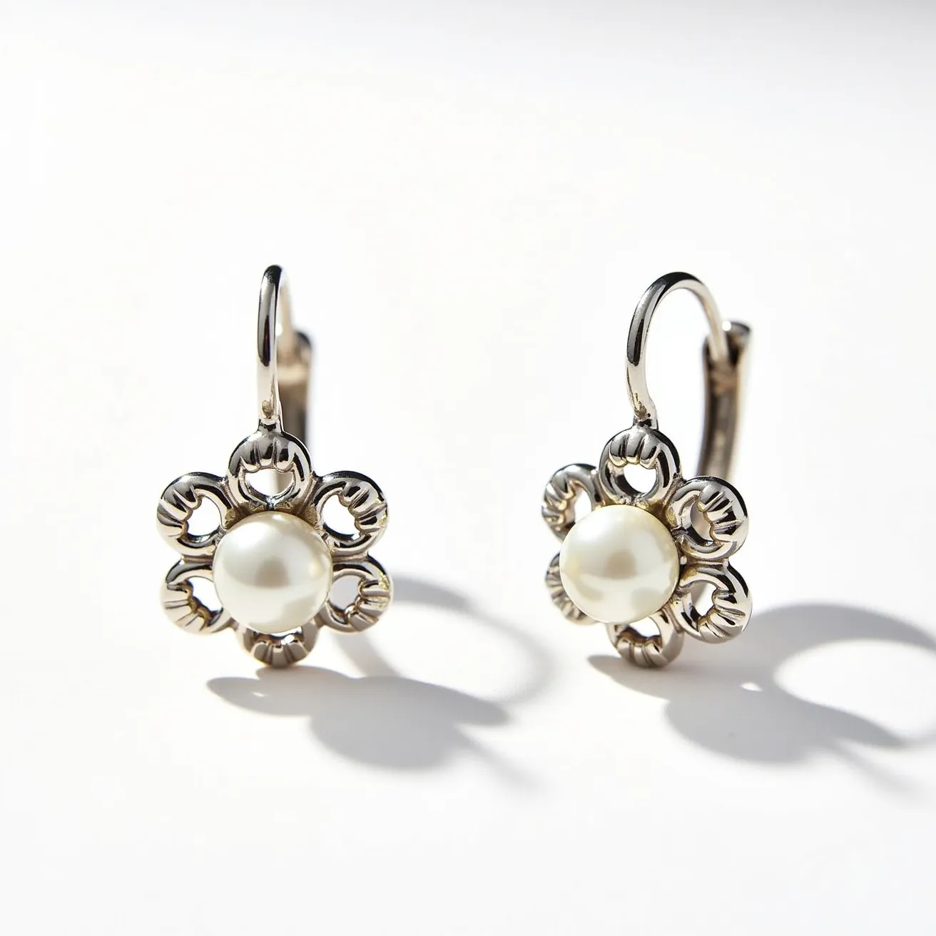 These leverback earrings feature a delicate and intricate design, crafted from a polished metal, possibly silver or white gold, forming a floral motif. At the center of each earring sits a luminous round pearl, which serves as the main gem, adding an elegant and classic touch. The pearls are held securely in place by the metal setting that mimics petal-like shapes around them. The leverback clasp ensures a secure and comfortable attachment to the ear, combining both sophistication and practical functionality.