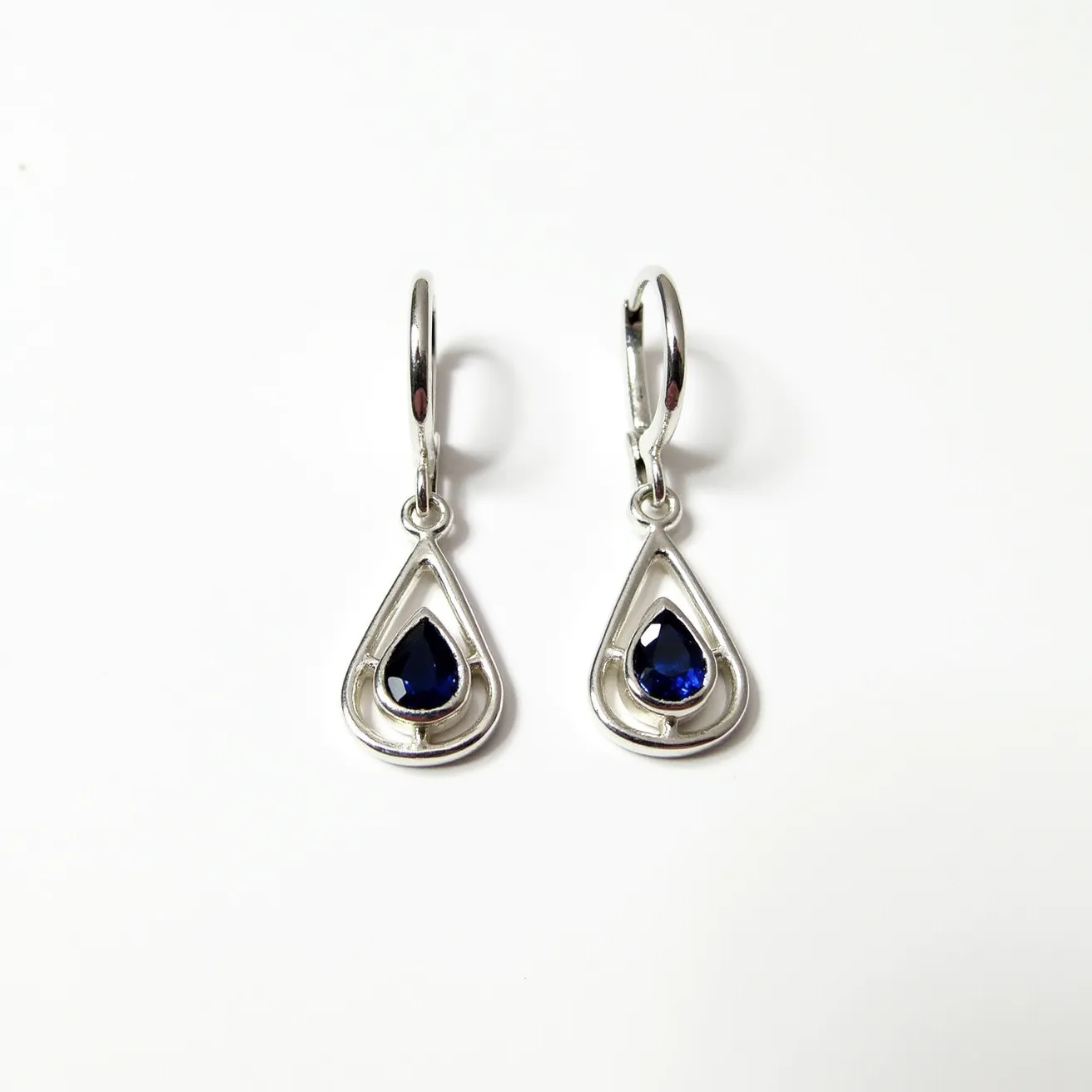 These leverback earrings feature an elegant design crafted from a polished silver material, showcasing a sophisticated and contemporary appearance. The focal point of each earring is a deep blue teardrop-shaped gemstone that is exquisitely cut to enhance its reflective qualities. These gemstones are securely set within a teardrop-shaped silver frame that complements their shape while adding a layer of protection and aesthetic appeal. The leverback clasp attaches the earrings securely, providing both ease of use and a stylish finish to the overall design.