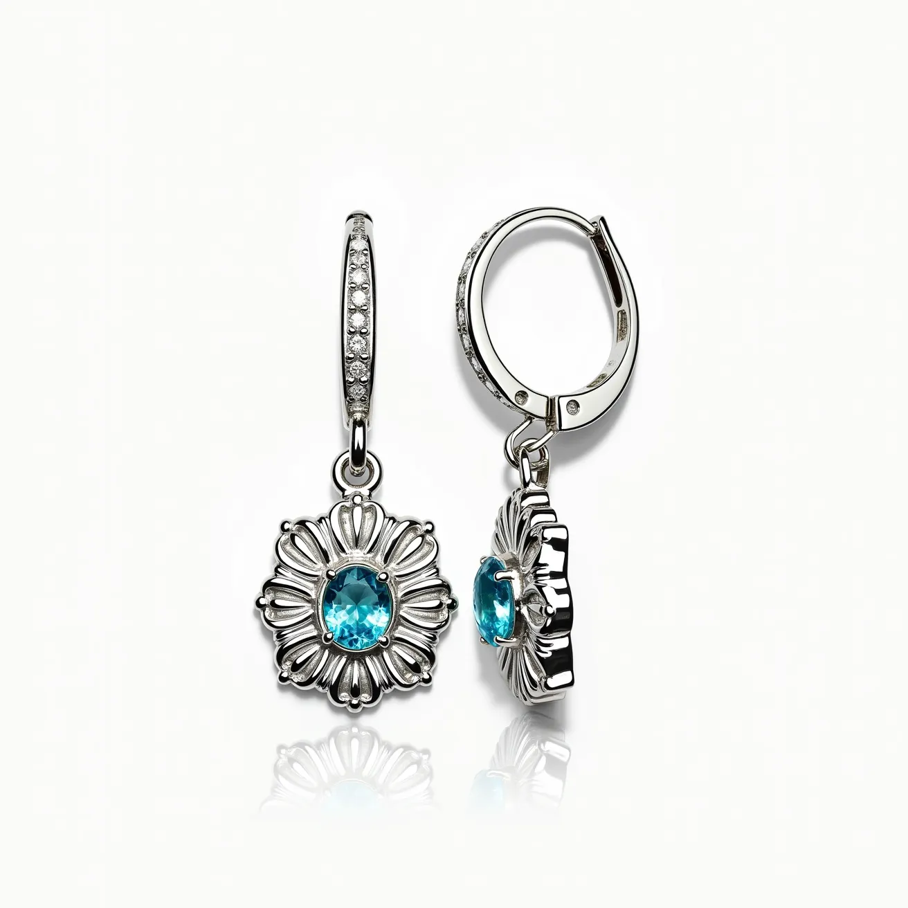 These little girl earrings feature an intricate design with a beautiful blue gemstone, likely a topaz, at their center. The stone is oval-cut and surrounded by a detailed, floral-like silver setting that adds elegance and charm. The hoops are adorned with tiny, sparkling clear stones, enhancing their overall brilliance. The earrings are equipped with a hinged hoop clasp, providing a secure and comfortable fit. The combination of the vibrant blue stone and the shimmering clear accents creates a delightful and eye-catching accessory.