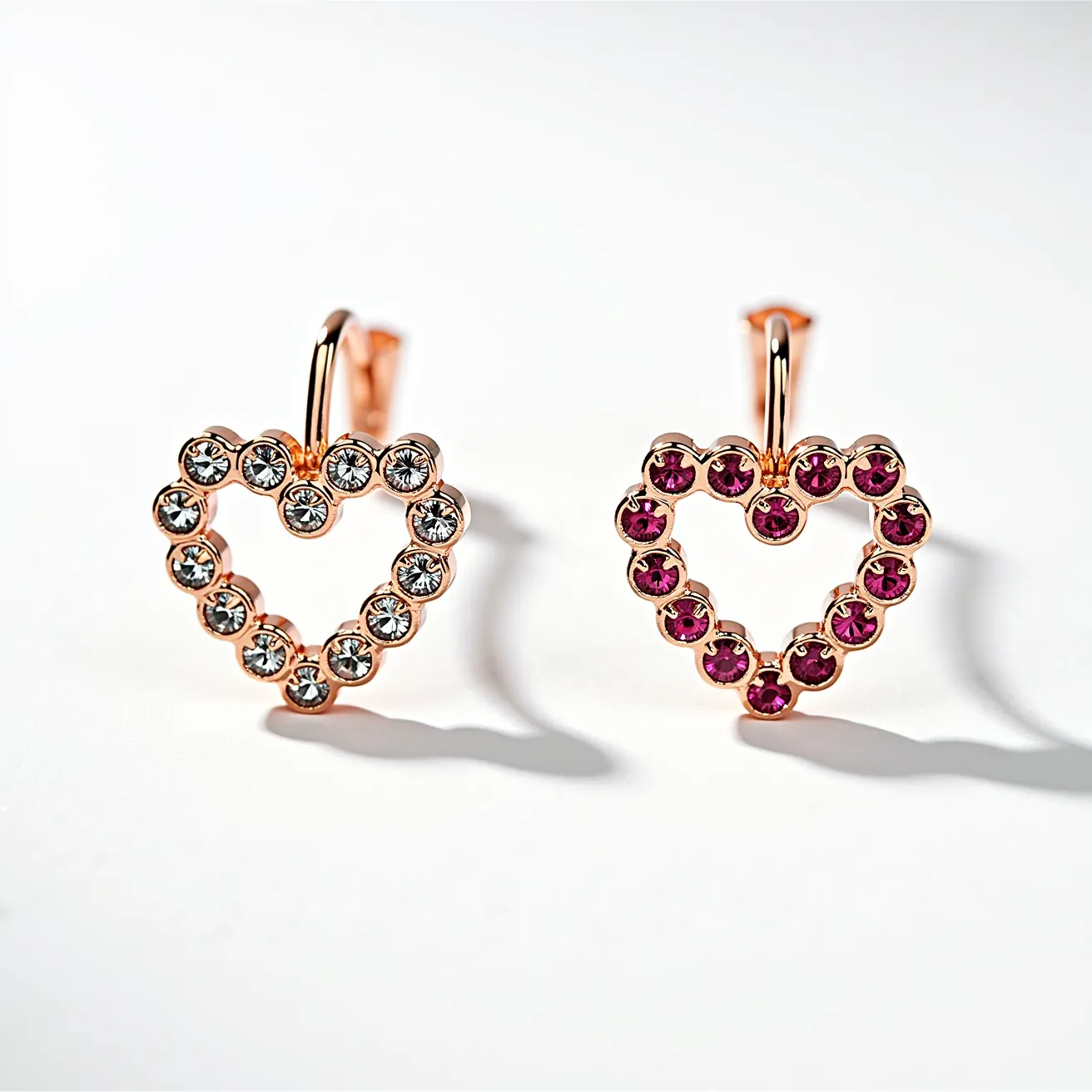 These little girl earrings feature two heart-shaped designs, each crafted with a rose gold metal base. The earrings are adorned with small, round gemstones set in a prong setting. One heart is lined with clear, sparkling stones that resemble diamonds or cubic zirconia, while the other is accented with vibrant pink stones, possibly rubies or pink sapphires. Each earring is equipped with a simple hook clasp for easy attachment. The overall design is delicate and youthful, perfectly suited for a little girl.