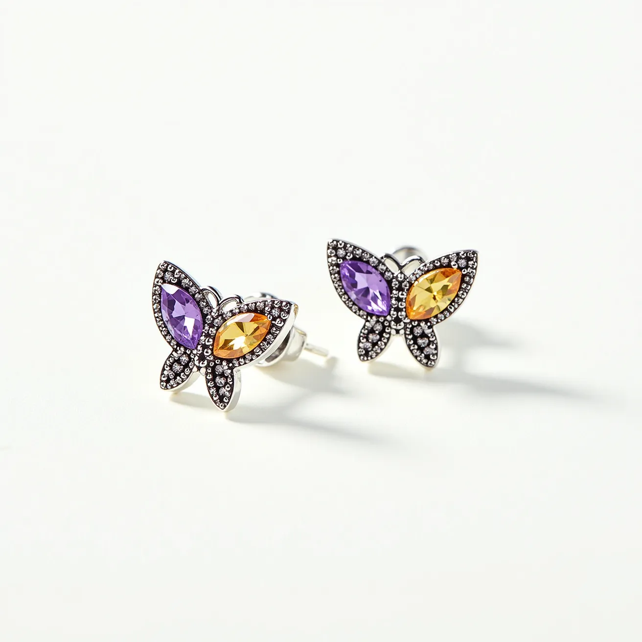 These little girl's earrings are designed in the shape of butterflies, showcasing a charming combination of vibrant gemstones. Each butterfly features two marquise-cut stones, one purple and one yellow, set in a delicate arrangement to form the wings. These stones are surrounded by a metal setting adorned with small, sparkling accents that resemble pavé or bead settings, enhancing the earrings' visual appeal. The metal appears to be a polished silver-tone material, providing a shiny and elegant backdrop for the colorful gems. The earrings use a post and butterfly clutch attachment, ensuring a secure and comfortable fit.