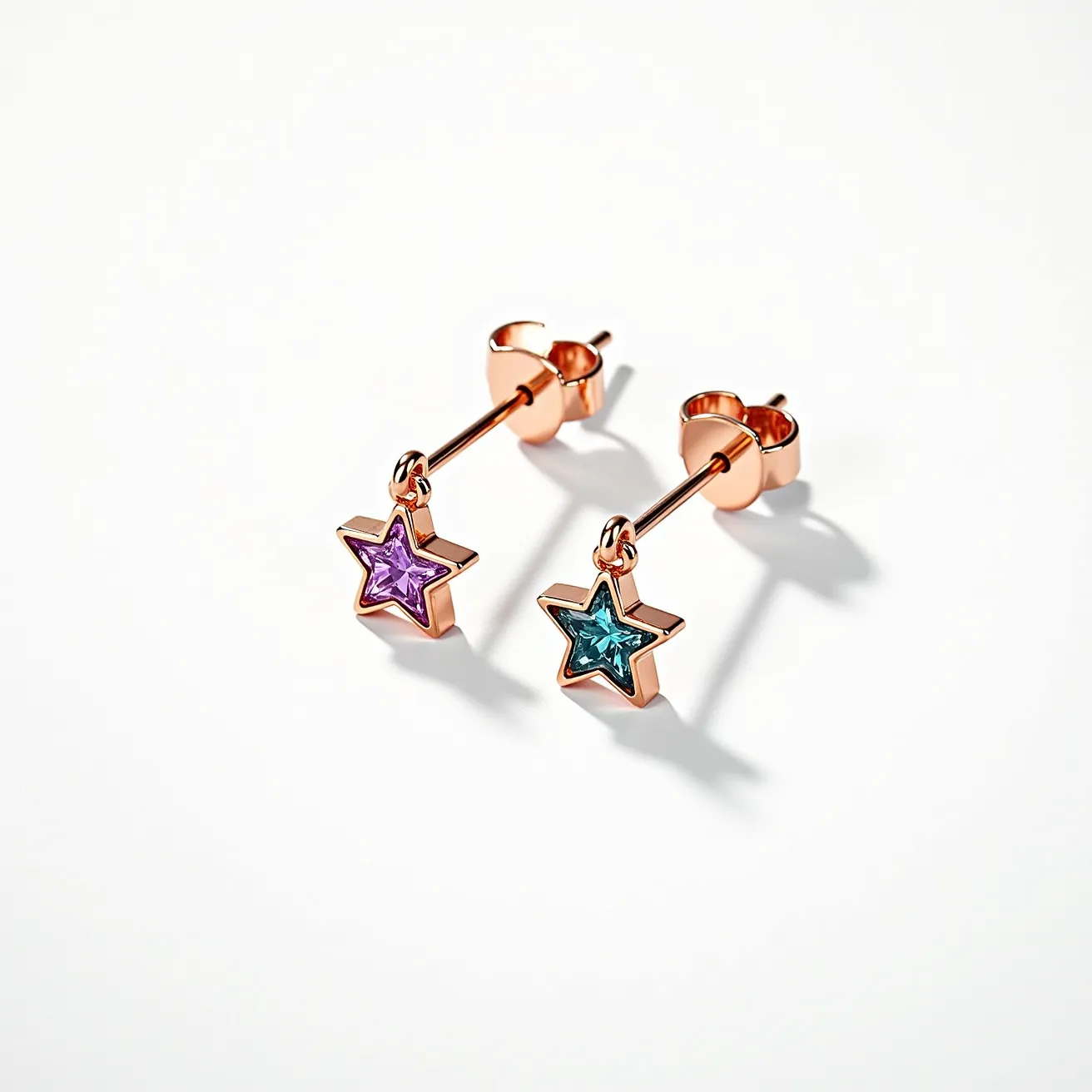 These little girl earrings feature charming star-shaped charms suspended from a stud post. The stars are adorned with vibrant gems—one in a deep purple and the other in teal. Each stone appears to be cut in a faceted style that enhances their sparkle and is set in a bezel setting, which securely encases the edges of each gem. The metal used for the earring structure and setting appears to be a rose gold tone, providing a warm contrast to the cool-colored stones. The attachment style is a standard post with a friction back, ensuring a secure and comfortable fit.