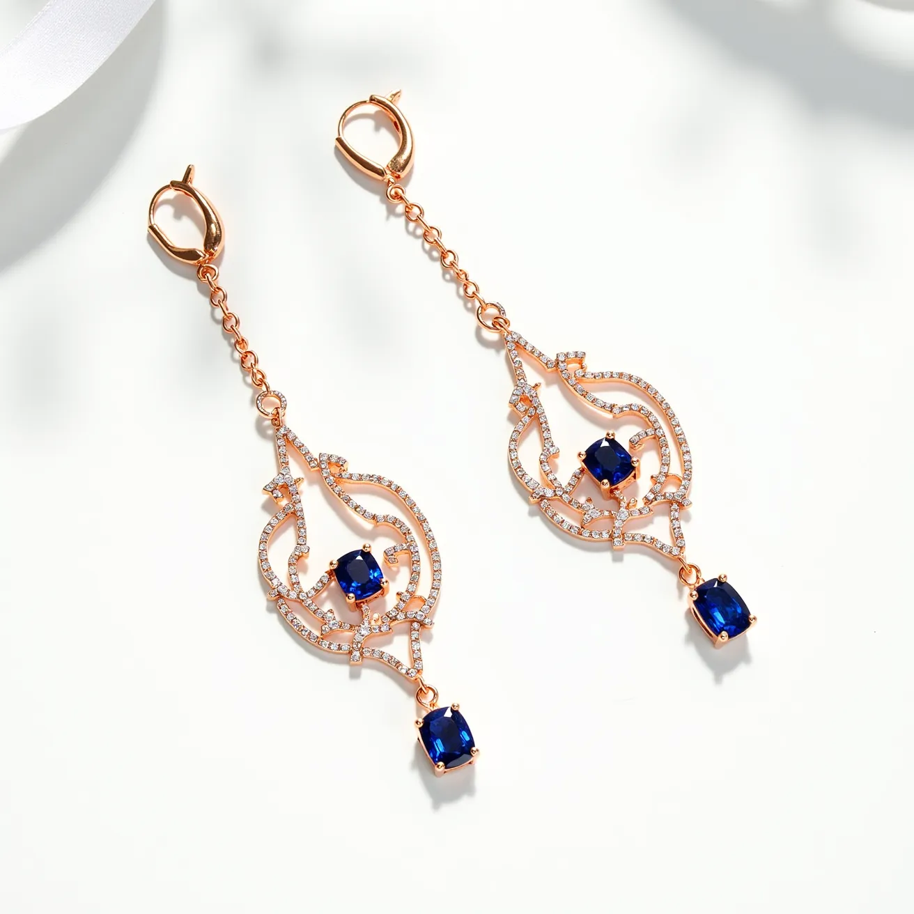 These long earrings showcase a sophisticated design featuring a base material in a lustrous rose gold tone. They are adorned with two dark blue, cushion-cut stones set in four-prong settings, which add an element of richness and depth. The earrings are further embellished with delicate, clear stones intricately arranged along the ornate framework, enhancing the overall elegance. The attachment piece is a small lever-back clasp, providing secure and comfortable wear. The intricate combination of the metallic swirl patterns with the vibrant blue stones and clear accents create a striking and luxurious accessory.