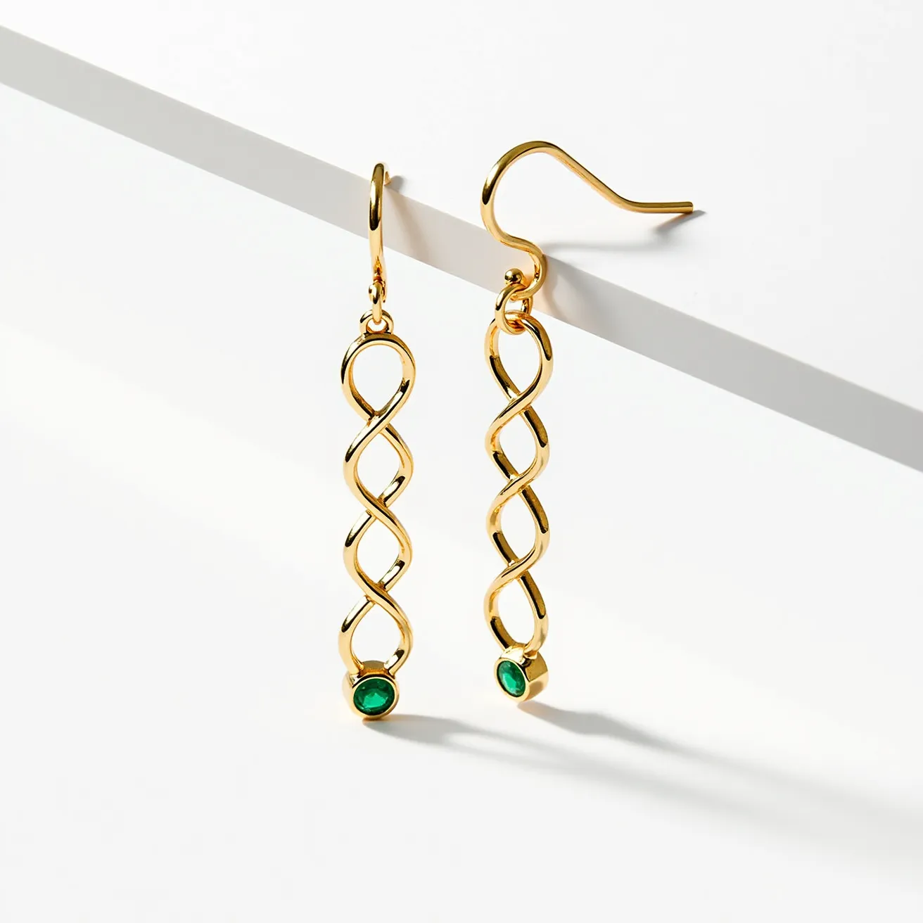 These long earrings feature an elegant, intertwined gold design that creates a visually appealing twisted pattern. At the base of each earring, there is a round green gemstone, likely an emerald, set into a simple bezel setting that highlights its vibrant color. The earrings are suspended from delicate hooks that allow for easy wear and movement, adding to their graceful and sophisticated appearance.