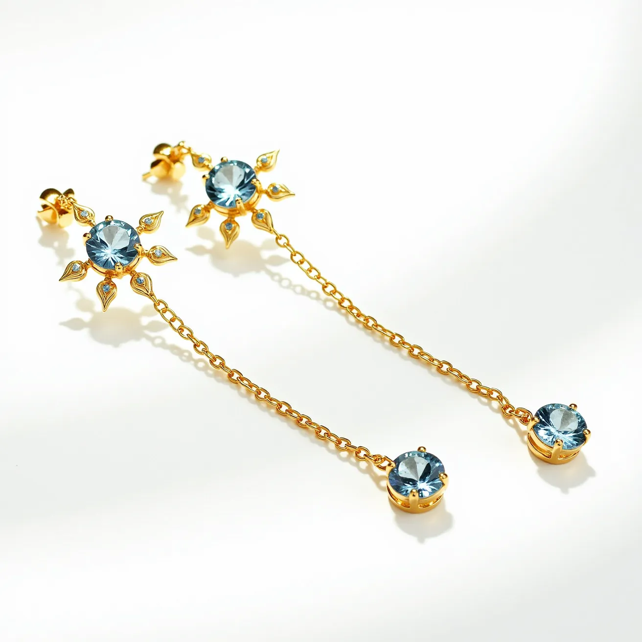 These long earrings feature a striking design with a gold chain that elegantly connects two main elements: a blue gemstone at the top and another at the bottom. Both stones are round-cut and appear to be set in a prong setting, which accentuates their brilliance. The upper gemstone is surrounded by a decorative pattern resembling sun rays, crafted from gold and enhanced with small, delicate leaf-like embellishments. The gold metal contrasts beautifully with the blue stones, creating a harmonious and eye-catching appearance. At the top, the earrings attach with a post and butterfly clutch, ensuring a secure fit.